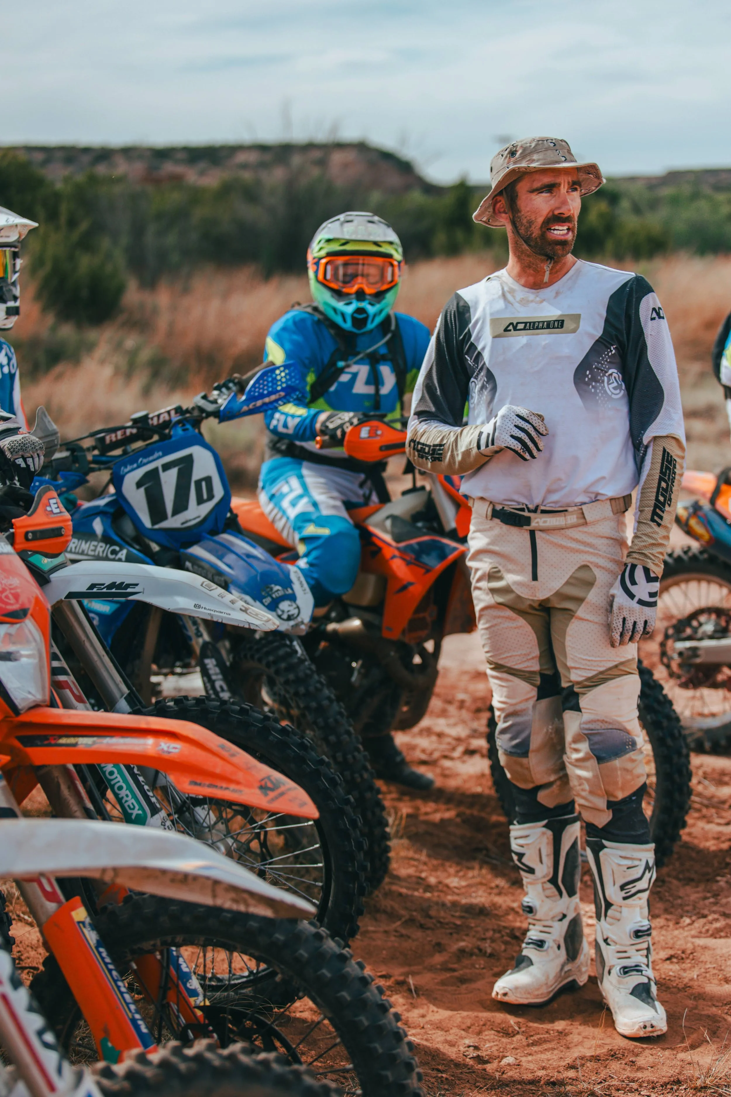 A man in motocross gear standing among dirt bikes outdoors, with another rider on a blue bike wearing a helmet and goggles in the background. The setting appears to be a dirt track or trail.