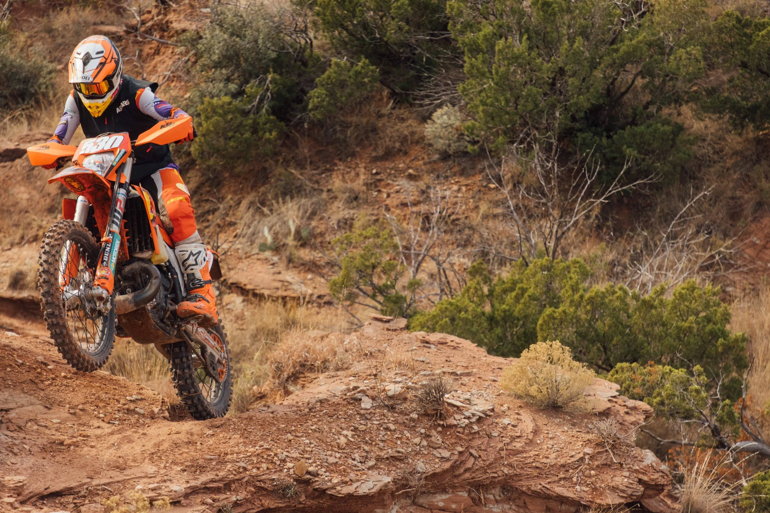 A person riding an orange dirt bike on a rugged trail in a desert landscape with sparse vegetation and bushes.