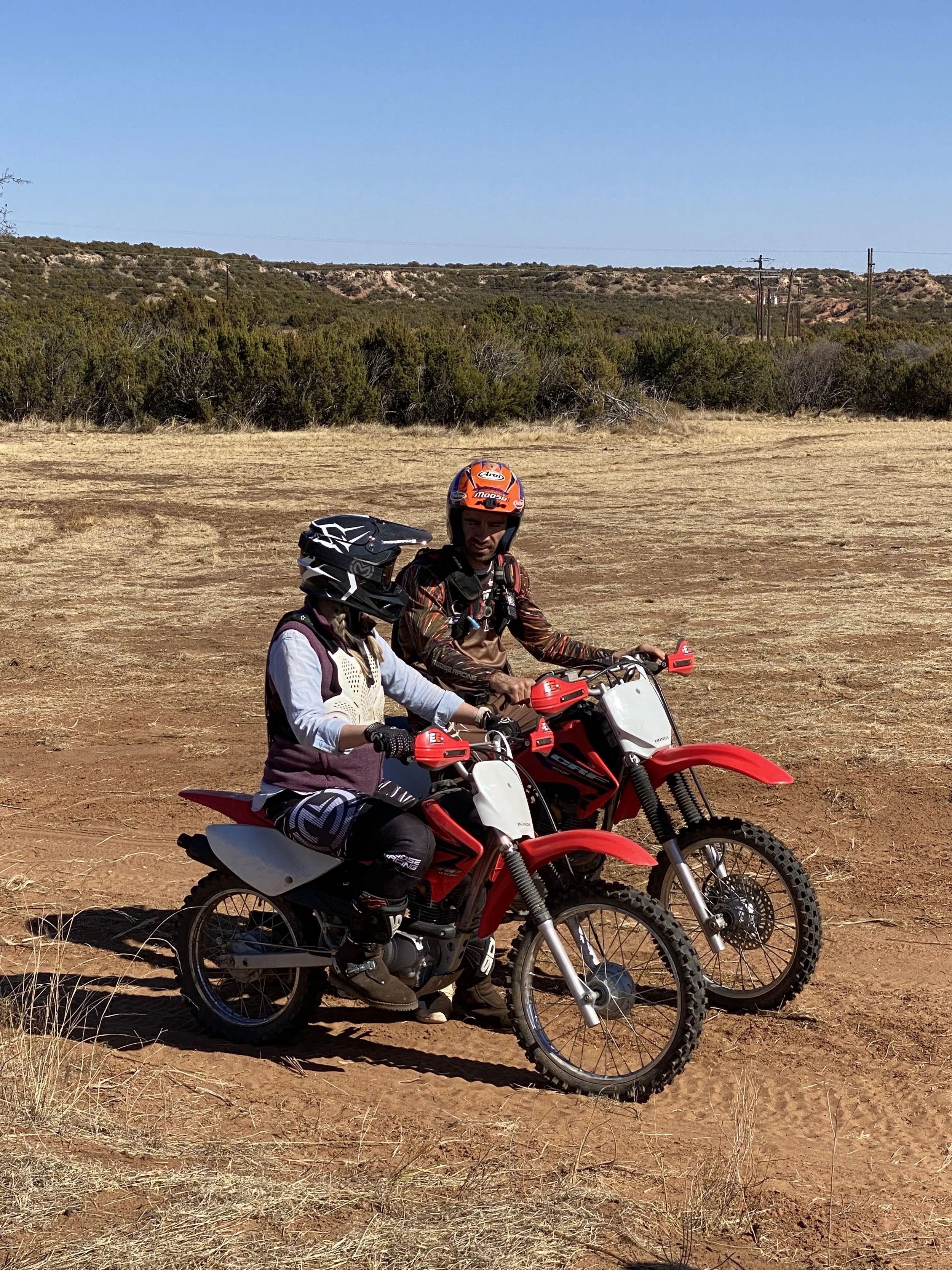 Two people in motocross gear and helmets sitting on red and black dirt bikes on a dirt field with shrubs and trees in the background under a clear blue sky.