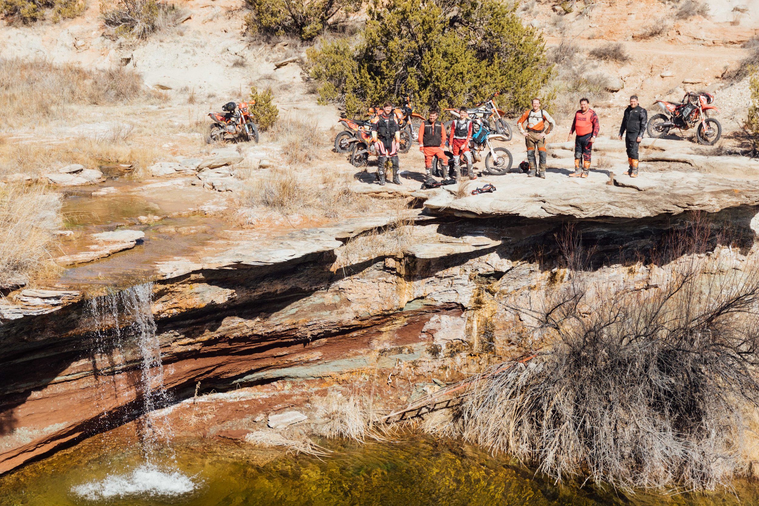 A group of six off-road motorcyclists standing with their dirt bikes in a desert landscape near a small waterfall and shallow stream, with sparse bushes and a large green bush in the background.