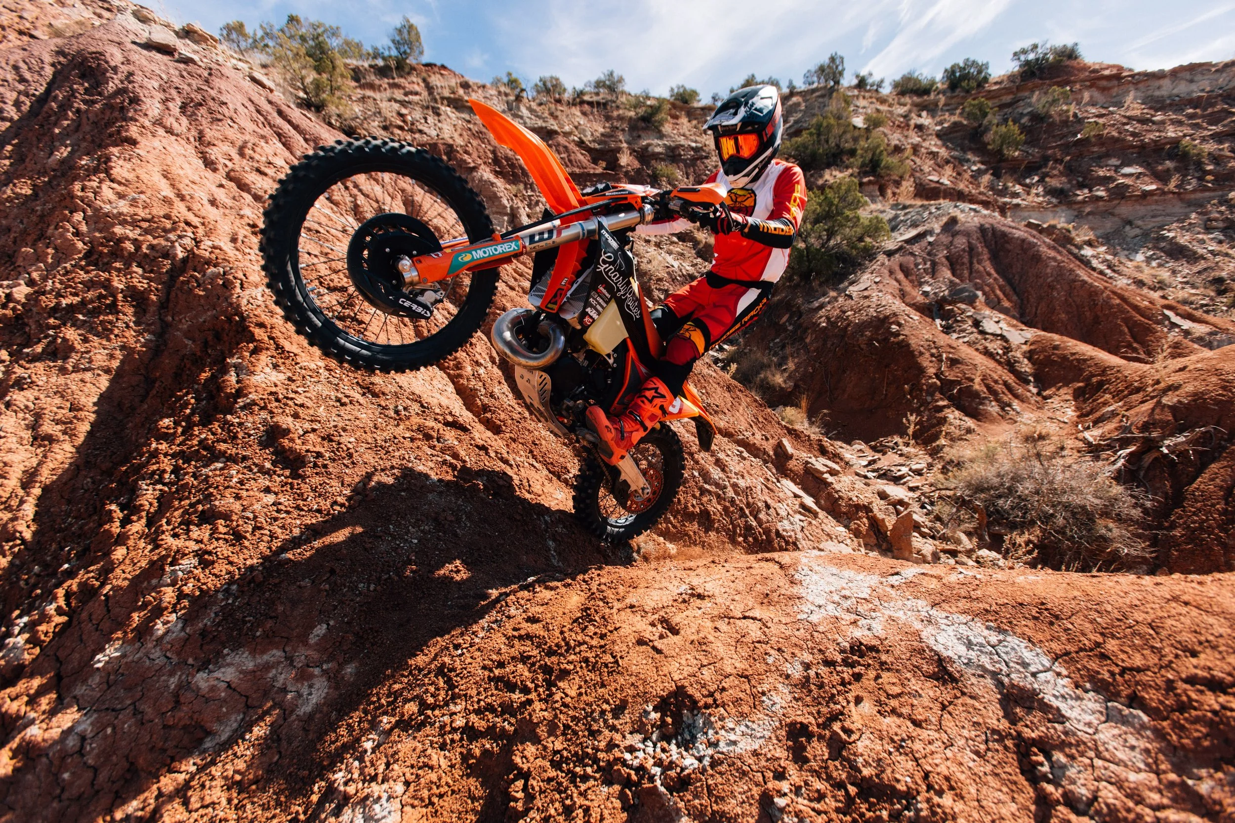 A person wearing a full-face helmet and red and white riding gear riding a dirt bike down a rocky, rugged terrain with reddish-brown soil and sparse desert vegetation.