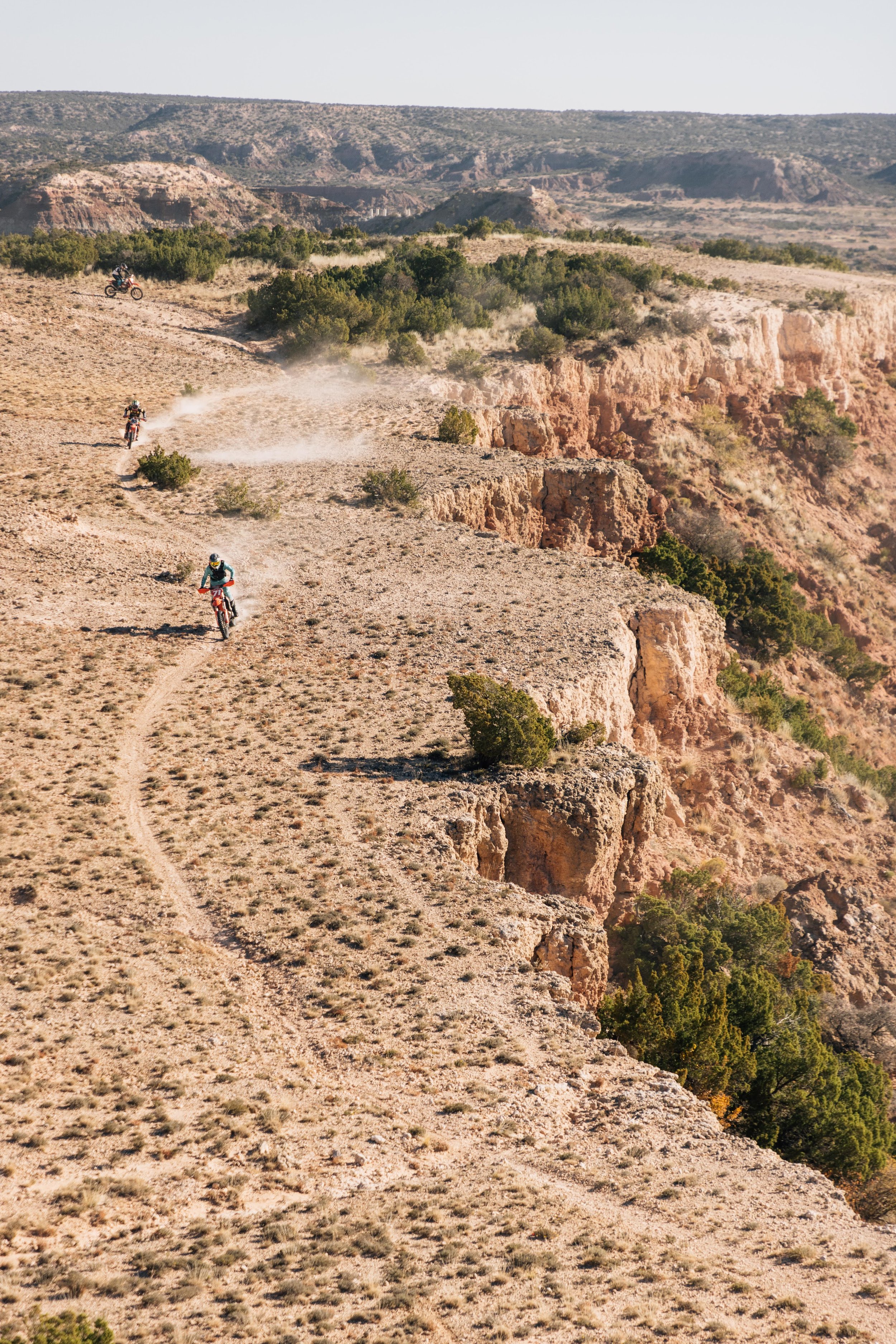 Four people riding dirt bikes on a rugged desert trail with rocky cliffs and sparse vegetation in the background.