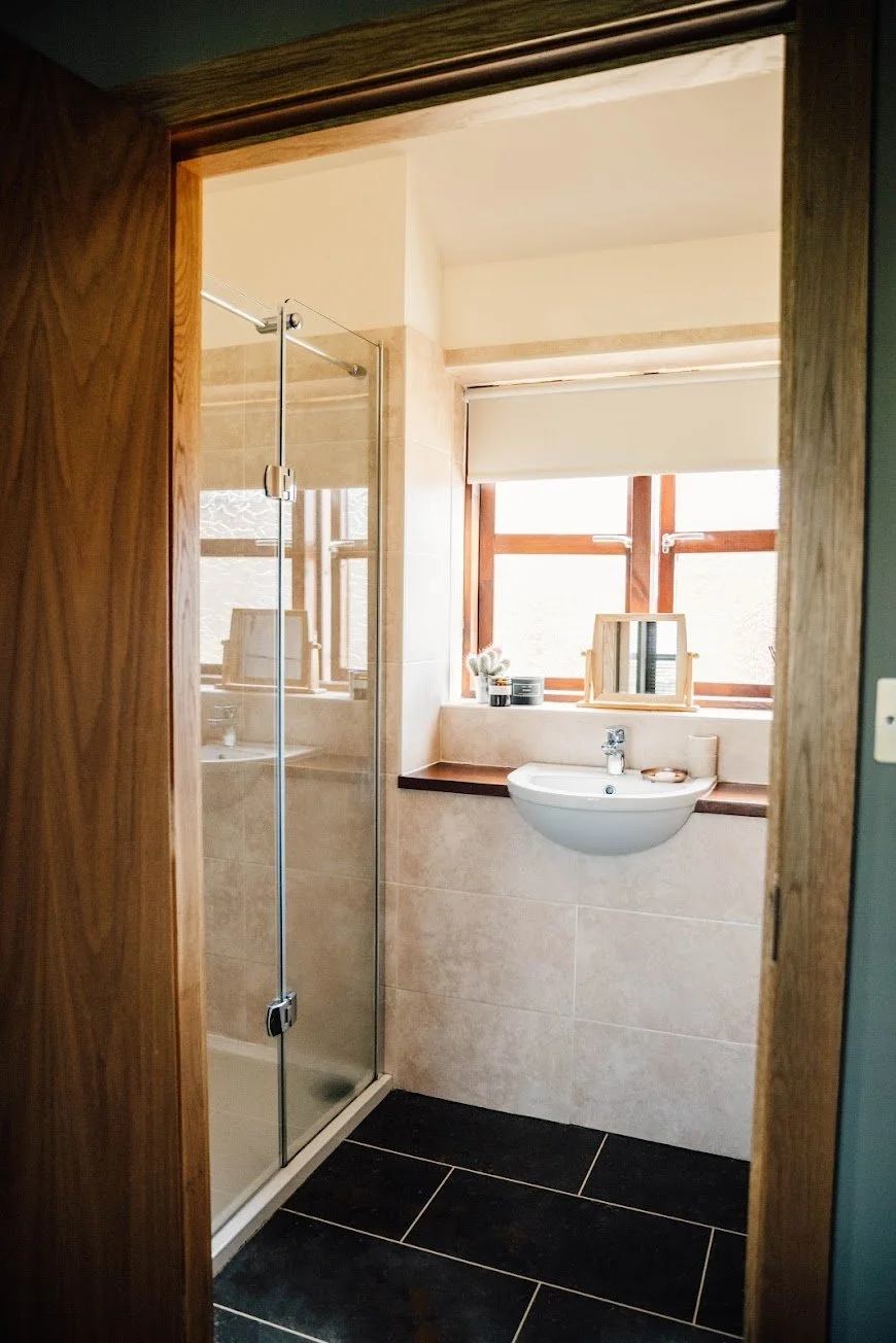 View into a small bathroom with a glass shower on the left, a wall-mounted sink beneath a window with a roller shade, and decorative items like framed mirrors and small containers on the window sill. The floor is tiled in dark color, and the walls are tiled in beige.