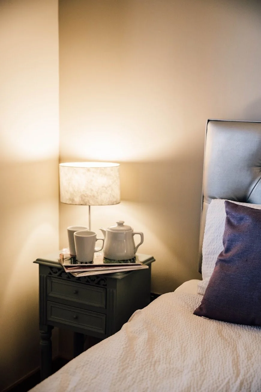 A cozy bedroom with a nightstand holding a lamp, a white teapot, and two cups, next to a bed with white bedding and purple pillows.