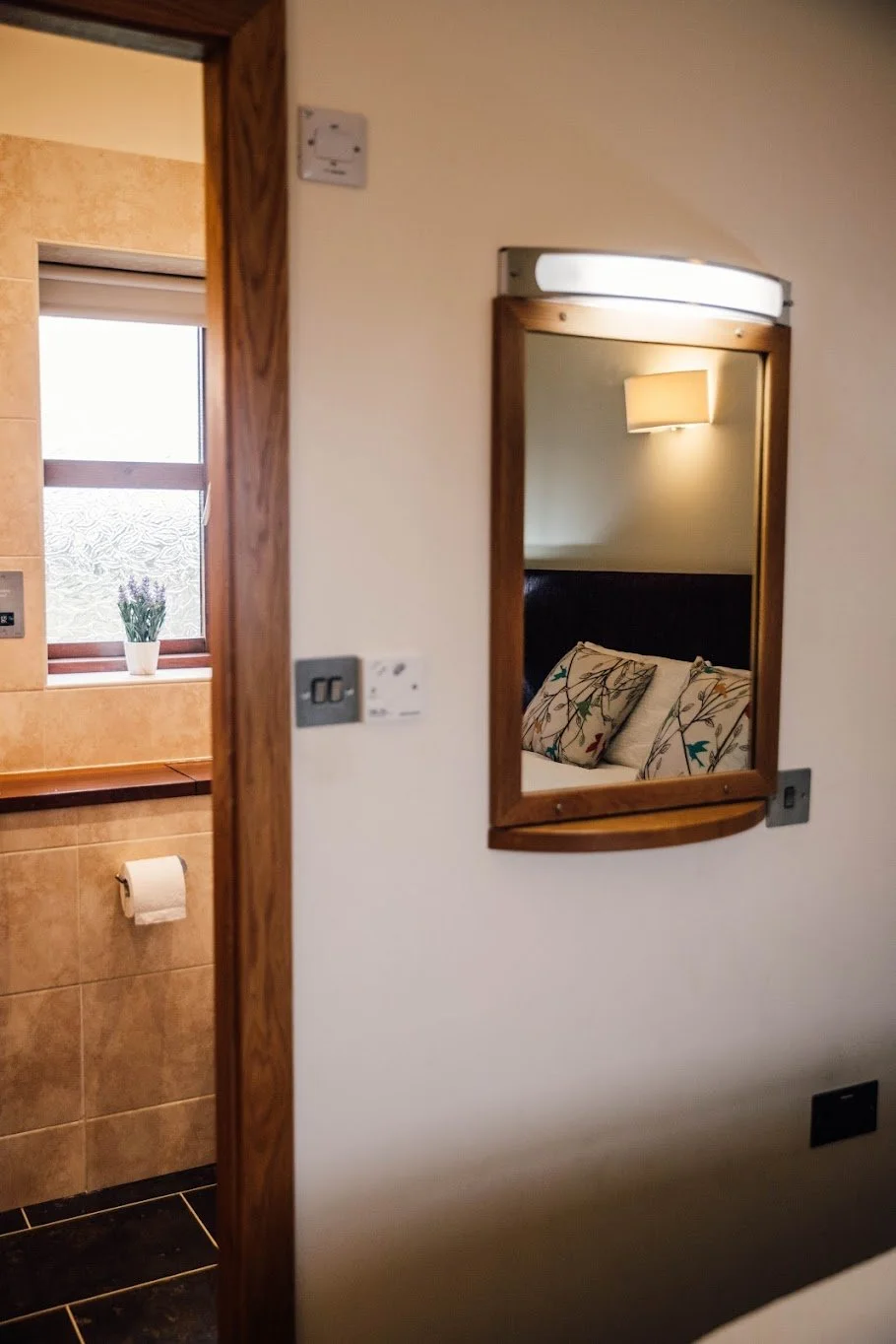 Reflection of a bedroom with a bed and floral pillows seen in a rectangular mirror on the wall. Part of a bathroom with a window, vase with flowers, and toilet paper holder is also visible.