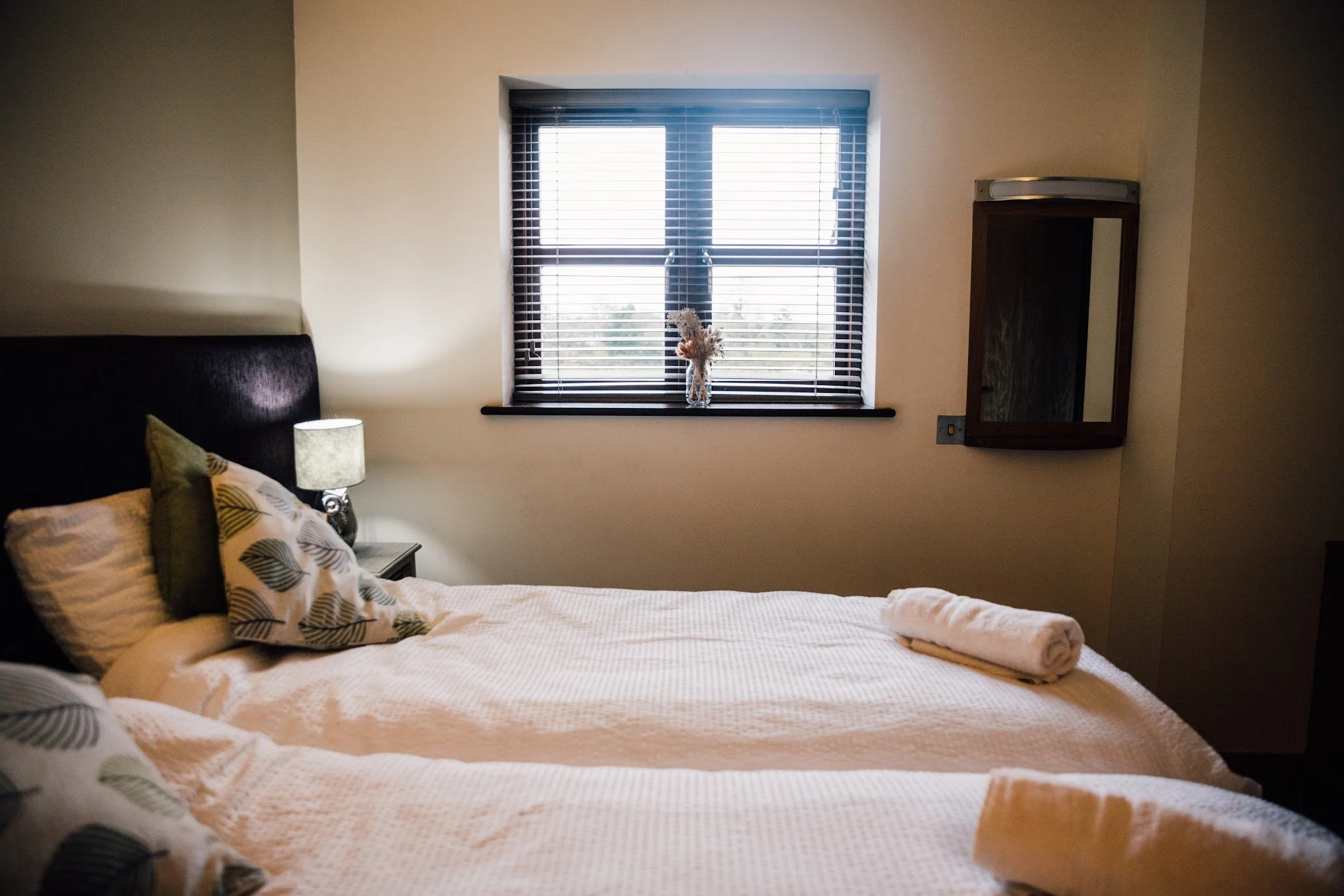A neatly made bed with a patterned pillow and a rolled towel, beside a nightstand with a lamp, in front of a window with blinds. A vase with dried flowers is on the windowsill, and a wall-mounted mirror is to the right of the window.
