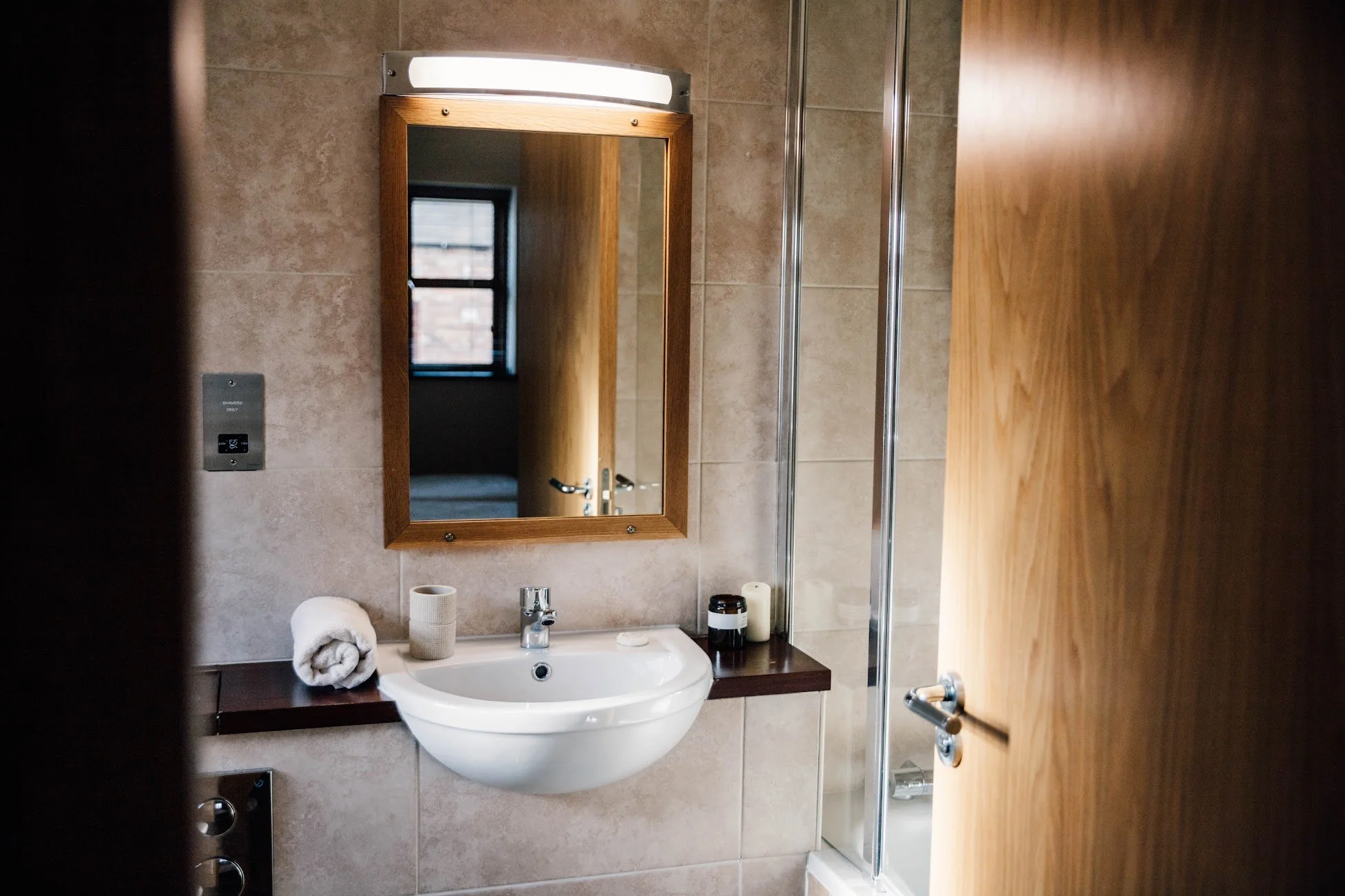 A bathroom sink with a cabinet, mirror, and wall-mounted light fixture. Items on the sink include a white rolled towel, a beige toothbrush holder, shampoo, and other toiletry bottles. The wall behind is tiled, and a window is reflected in the mirror.