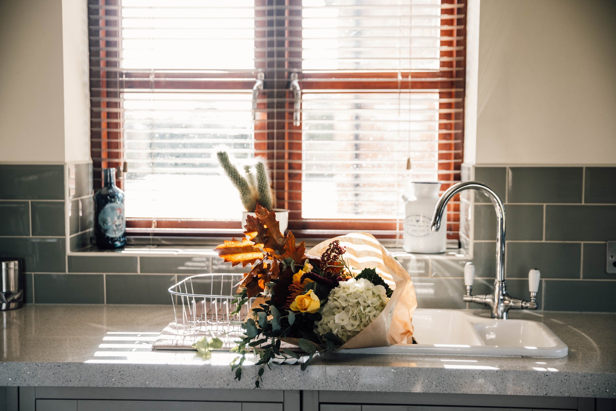 A kitchen sink with a bouquet of flowers and autumn leaves on the counter in front of a window with wooden blinds, sunlight streaming through.
