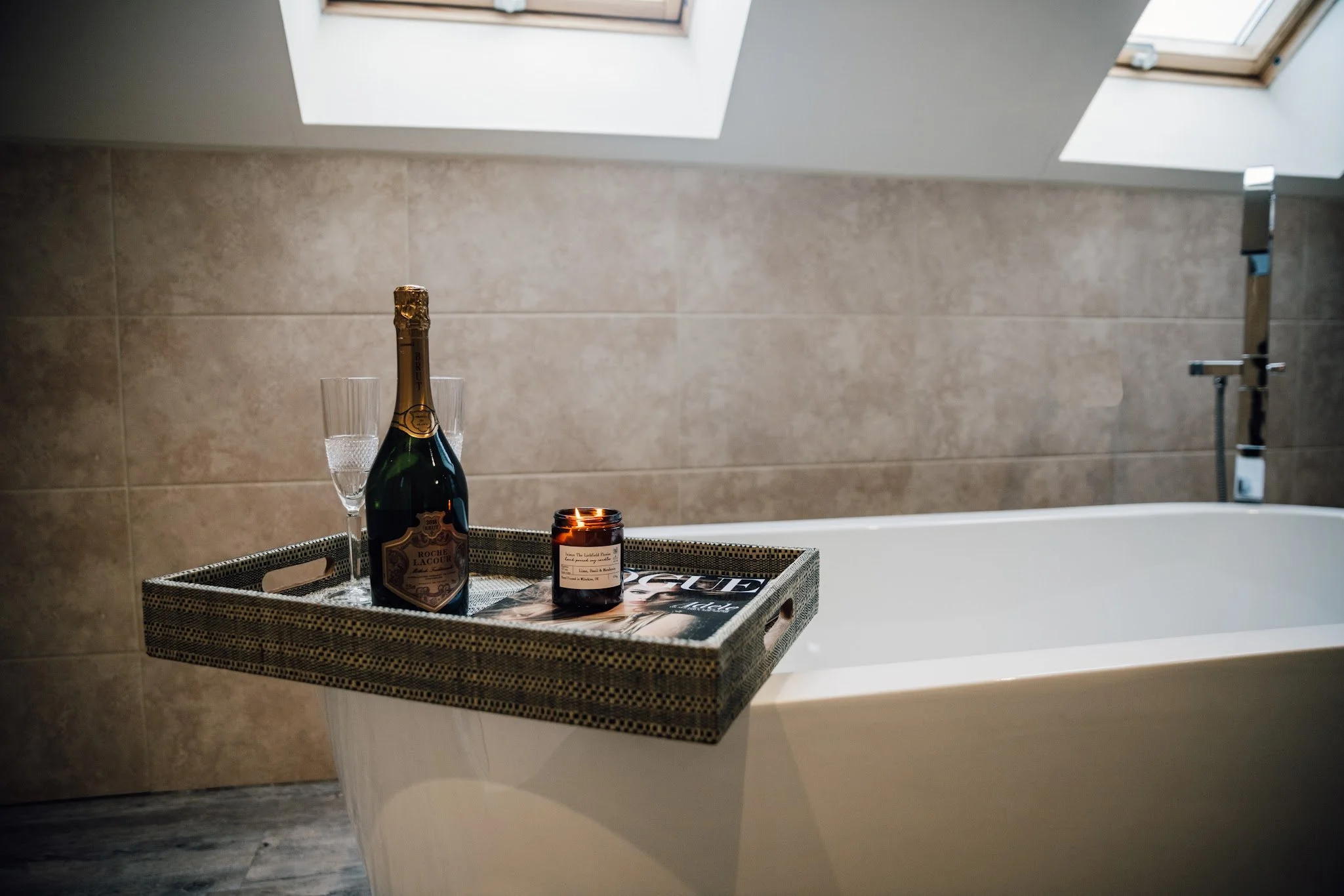 A bathtub with a showerhead in a bathroom with beige tiled wall and ceiling windows. A wicker tray on the edge of the tub holds a bottle of champagne, two champagne glasses, a lit candle, and a magazine.