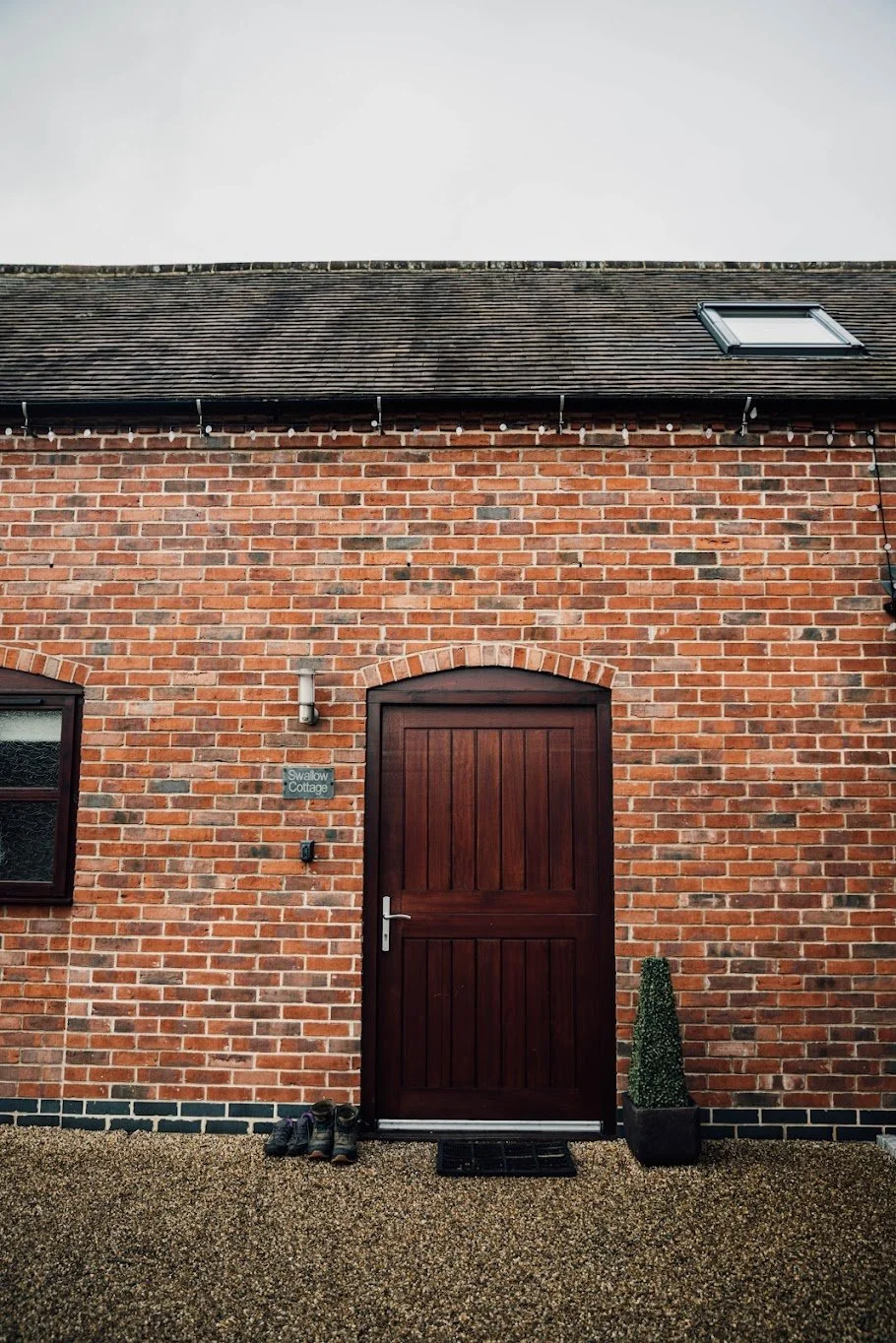 Brick house with a wooden door, a potted plant, shoes outside, a small window, and a sign that reads 'Swallow Cottage.'