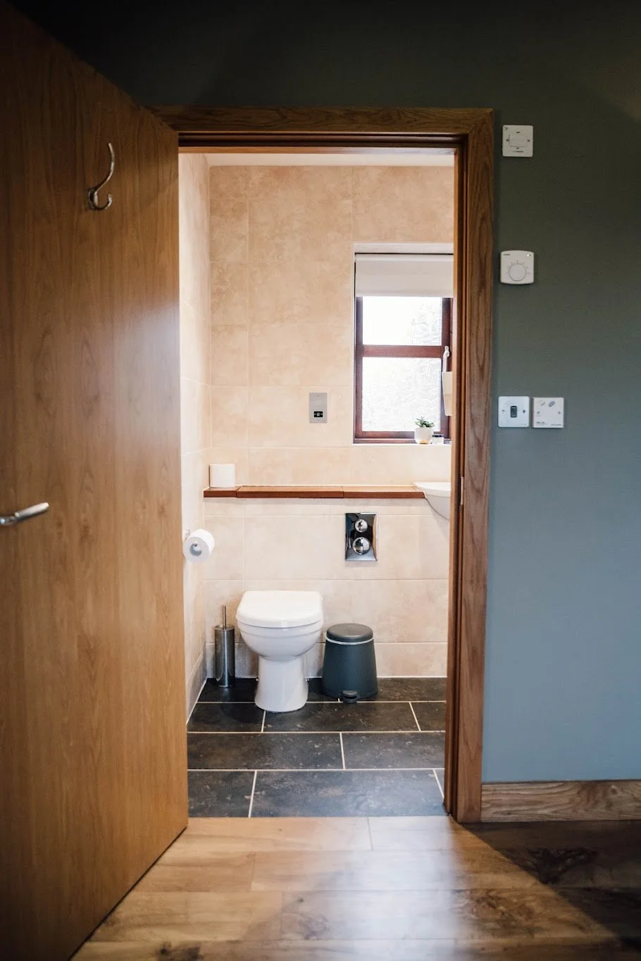 View into a small bathroom with a toilet, window, and a small plant. The bathroom has beige tiled walls and dark flooring, with a wooden doorframe.