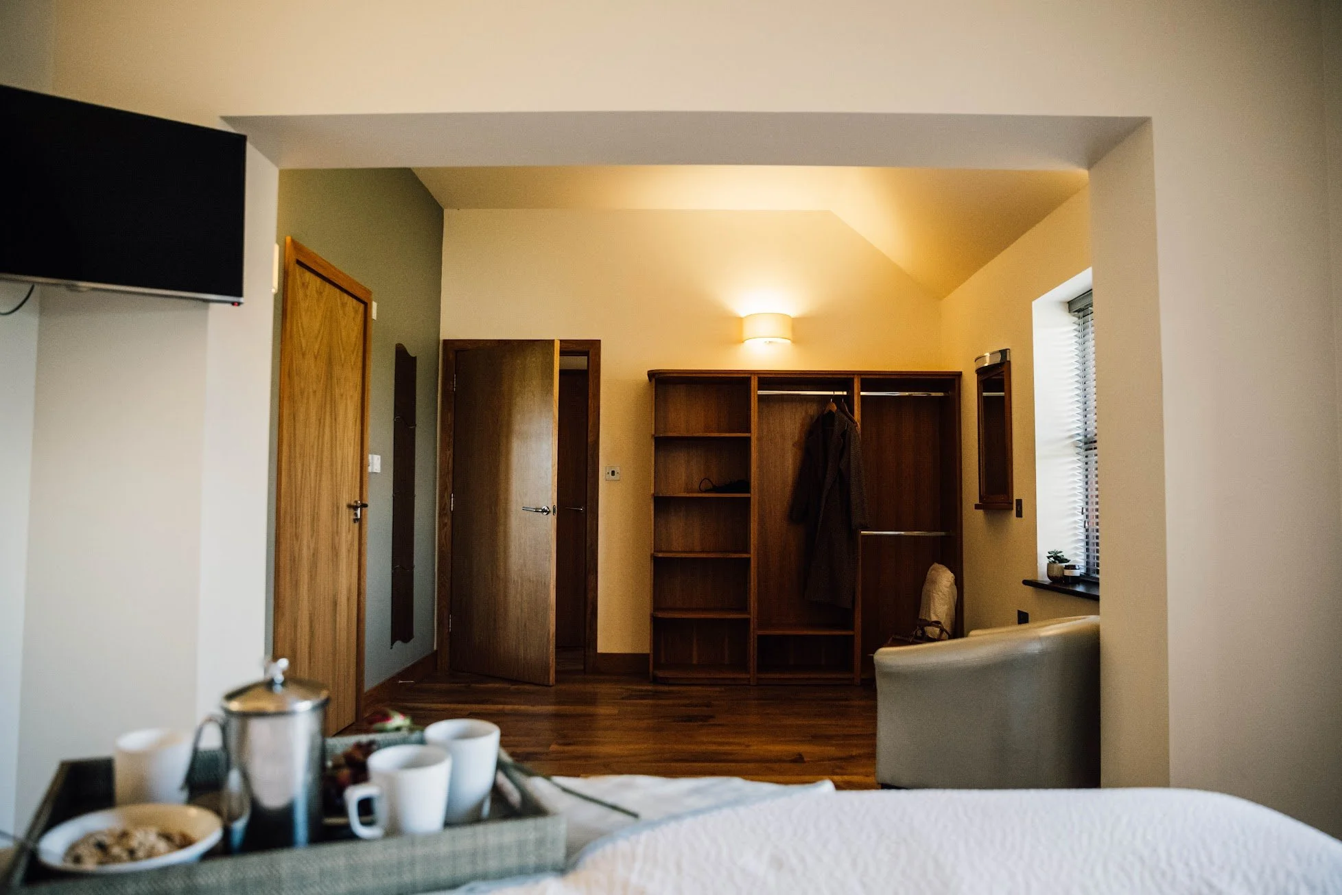 A cozy hotel room with a wooden wardrobe, a small window with blinds, a bed in the foreground with breakfast items on a tray.