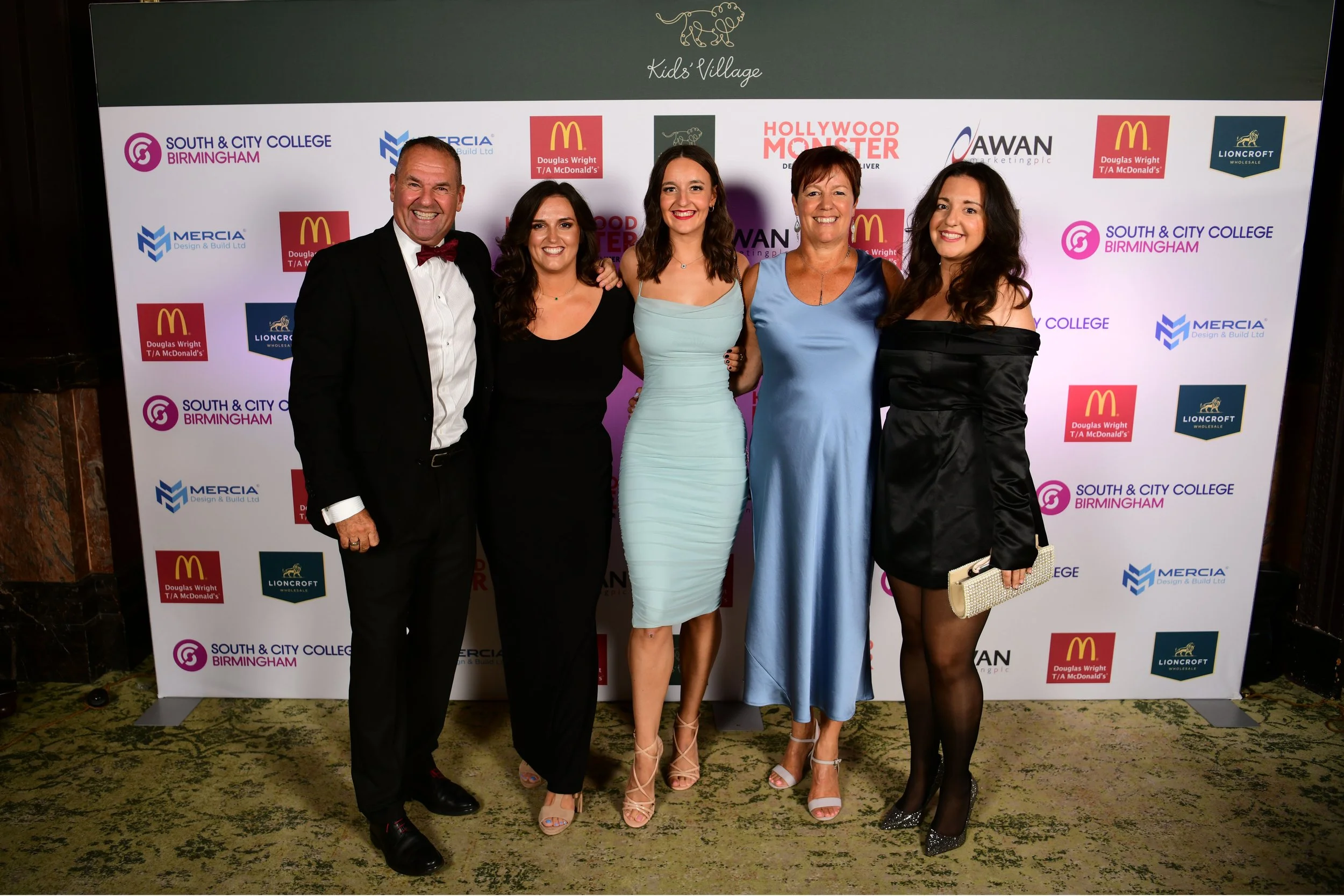 A group of five people, three women and two men, dressed formally, standing together and smiling at a photo event. They are in front of a step and repeat banner displaying logos for South & City College Birmingham, Mercia Design & Build Ltd, McDonald's, Lioncroft Wholesale, Hollywood Monster, and Awan Marketing.