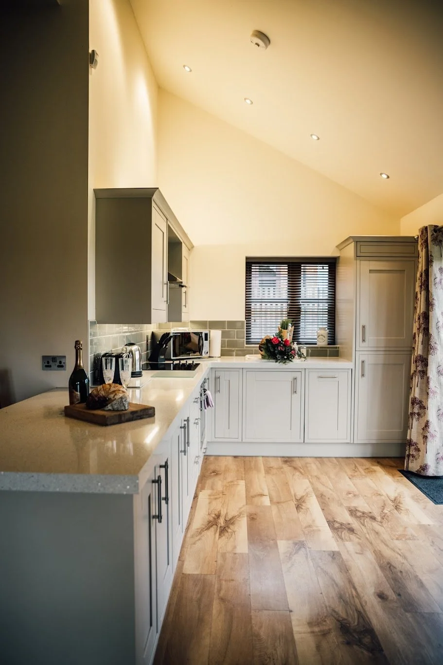 A cozy kitchen with white cabinets, wooden flooring, a window with blinds, a bouquet of flowers on the counter, and a floral curtain to the right.