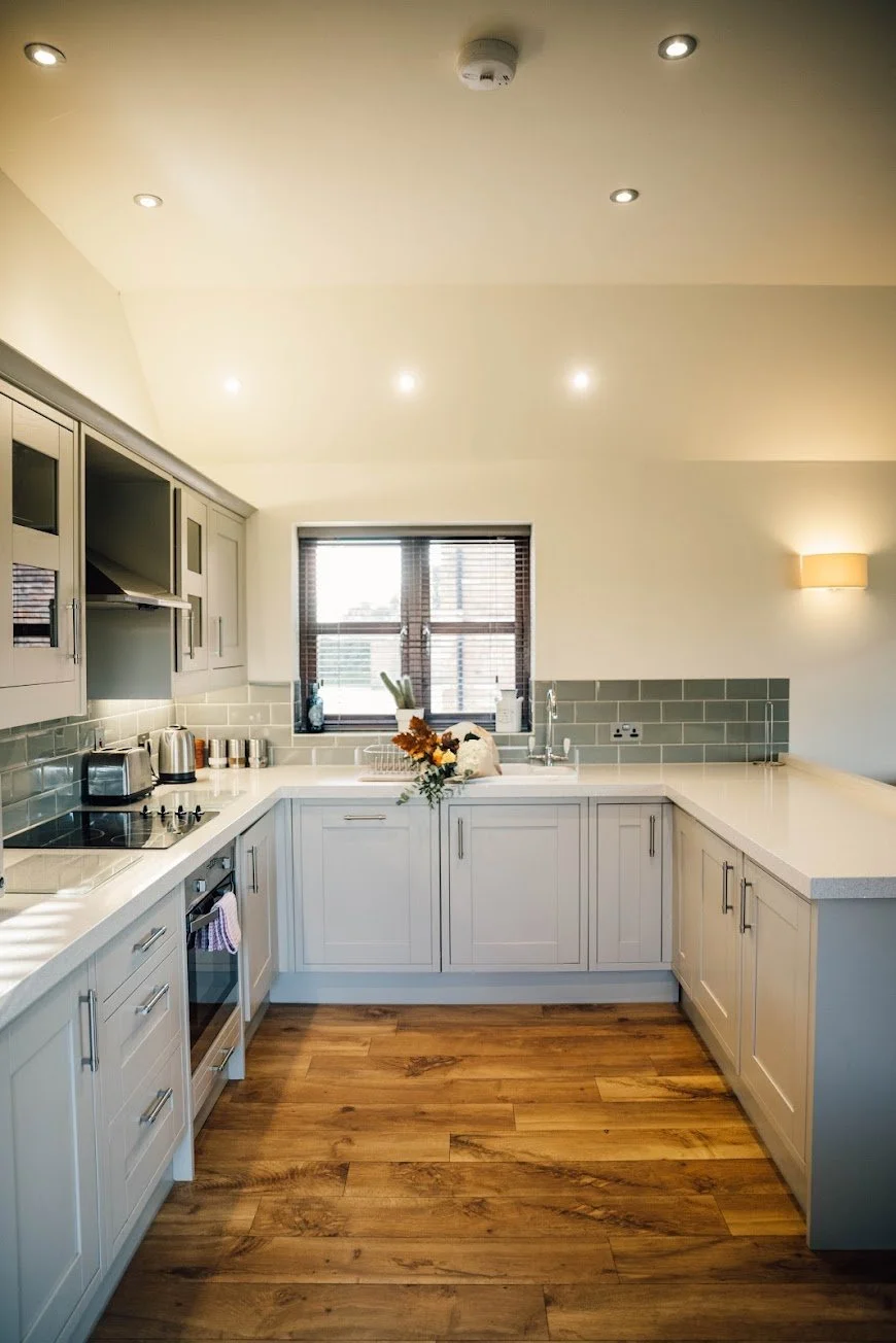 Modern kitchen with white cabinetry, wooden floors, a window with blinds, and a countertop with a bouquet of flowers and small appliances.