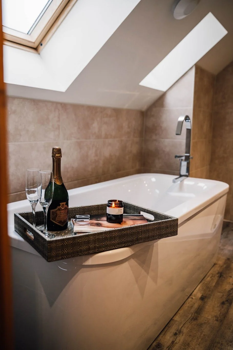 Bathtub with a tray holding champagne, glasses, and a lit candle in a cozy bathroom with a skylight window and wooden flooring.