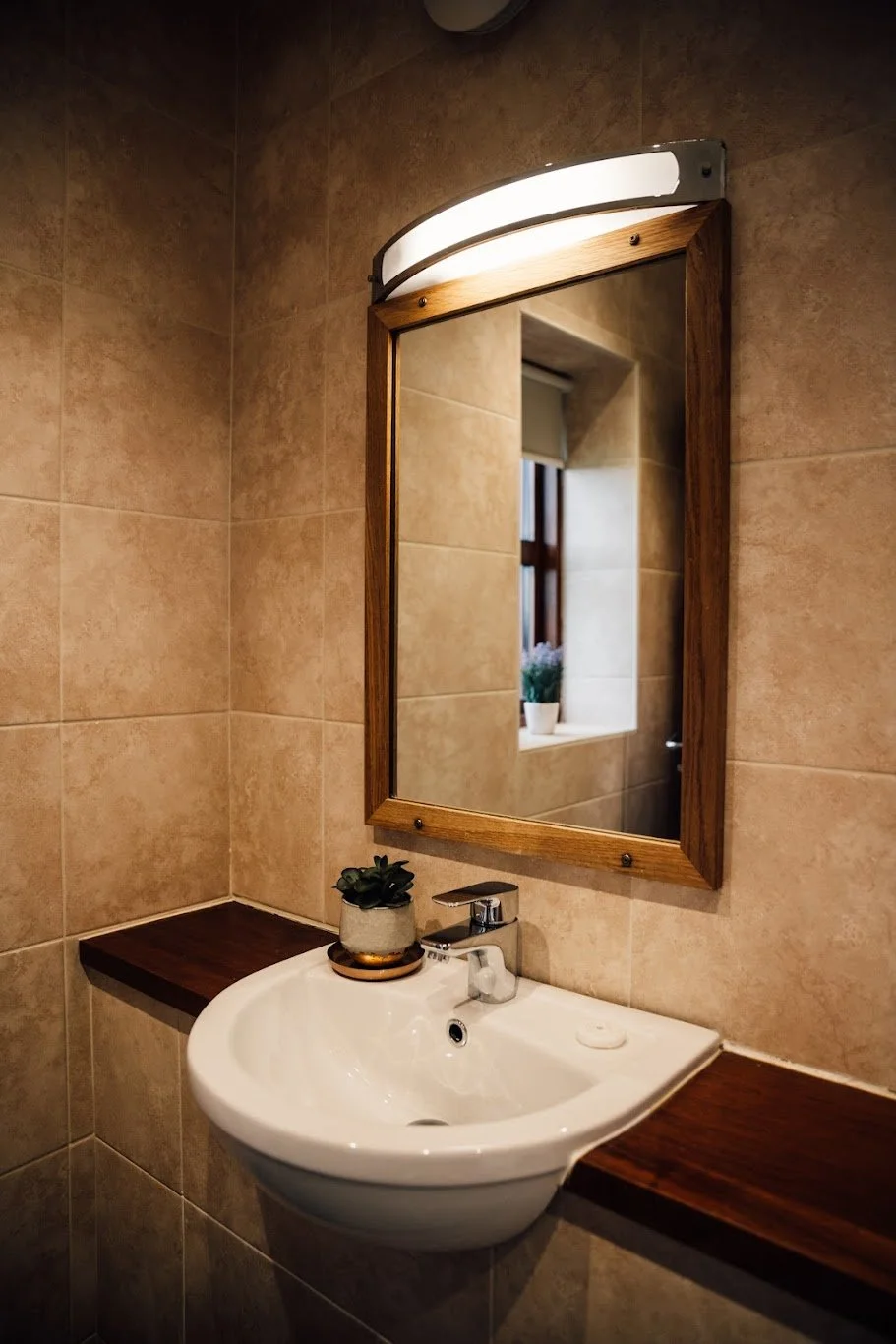 A bathroom with beige tiled walls, a white sink, and a wooden-framed mirror above it. There is a small plant on the sink and a window reflected in the mirror, with a potted plant on the window sill.