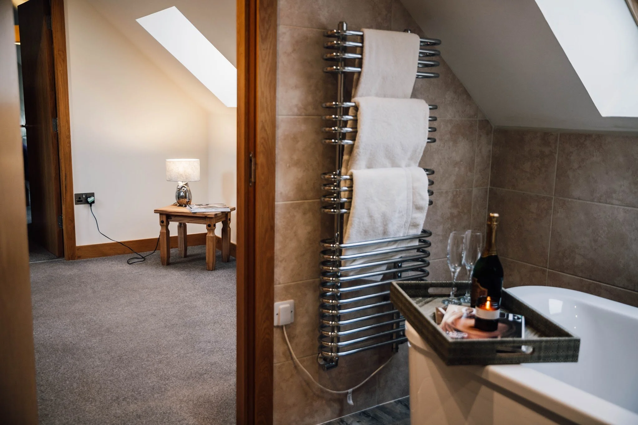 A bathroom with beige tiled walls and a chrome towel radiator holding two white towels. In the foreground, there is a vanity with a black tray holding a lit candle and some items, and two glasses. The background shows a doorway leading to a cozy room with a lamp on a wooden side table.