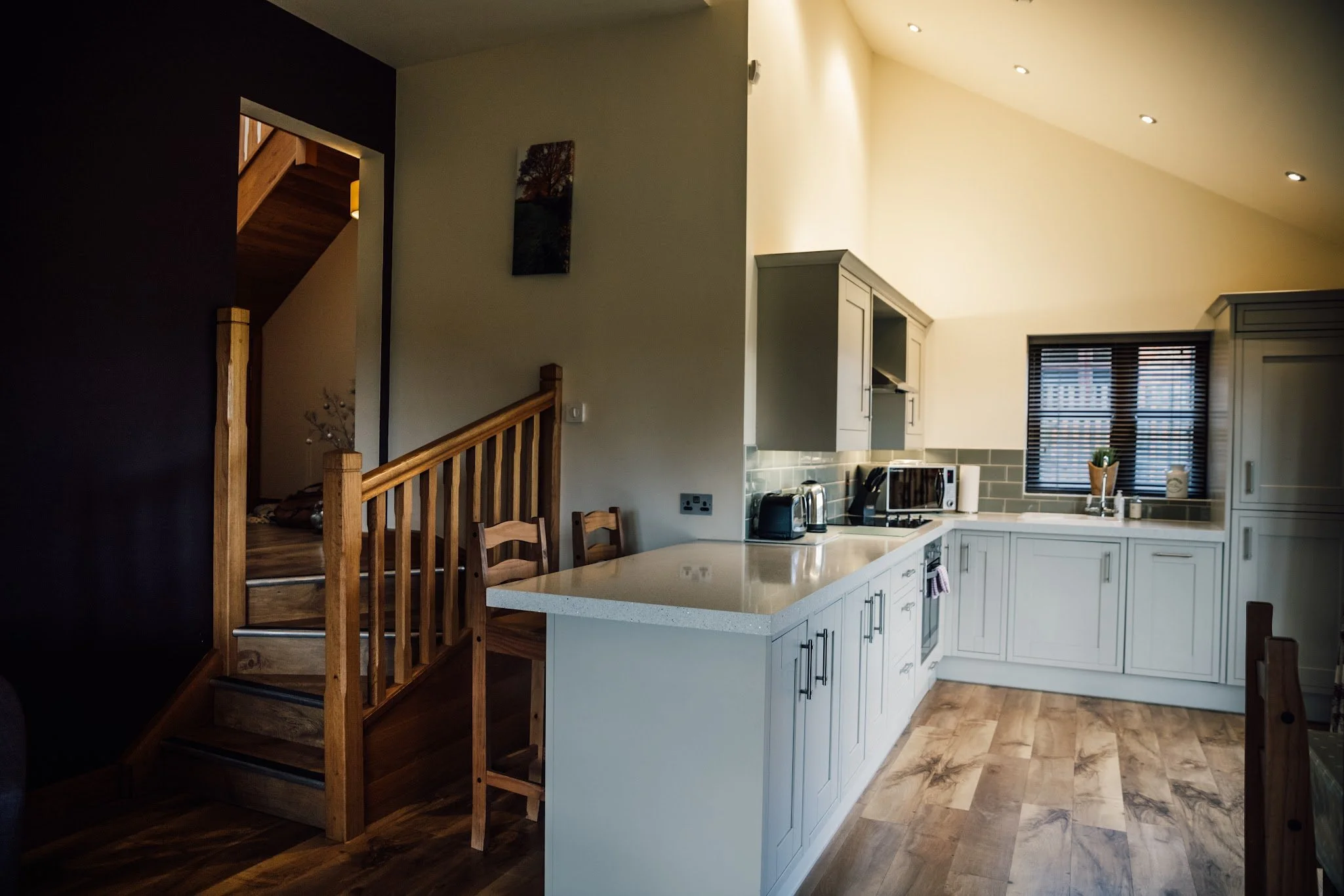 Inside a modern kitchen with white cabinets, a countertop with small appliances, and a window with blinds. Part of a wooden staircase and a black wall are also visible.