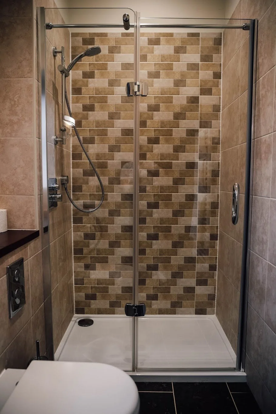 A modern walk-in shower with a glass door, beige and brown tile walls, a handheld showerhead, and a white shower base in a bathroom.