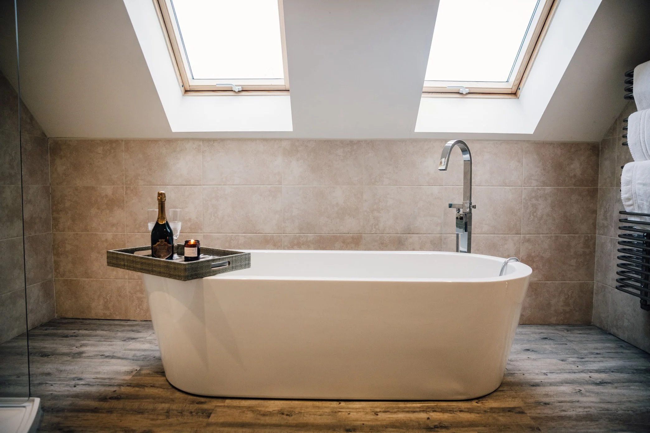 A modern bathroom with a freestanding bathtub under skylights. There is a tray with a bottle of champagne, two glasses, and a candle on the edge of the tub. Towel racks with white towels are mounted on the wall to the right.