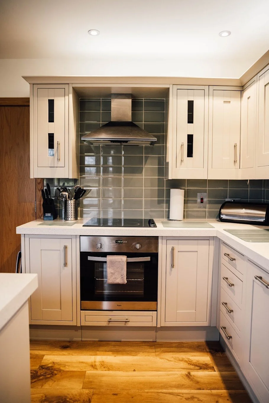 Modern kitchen with white cabinets, stainless steel oven and range hood, green tiled backsplash, and wooden flooring.