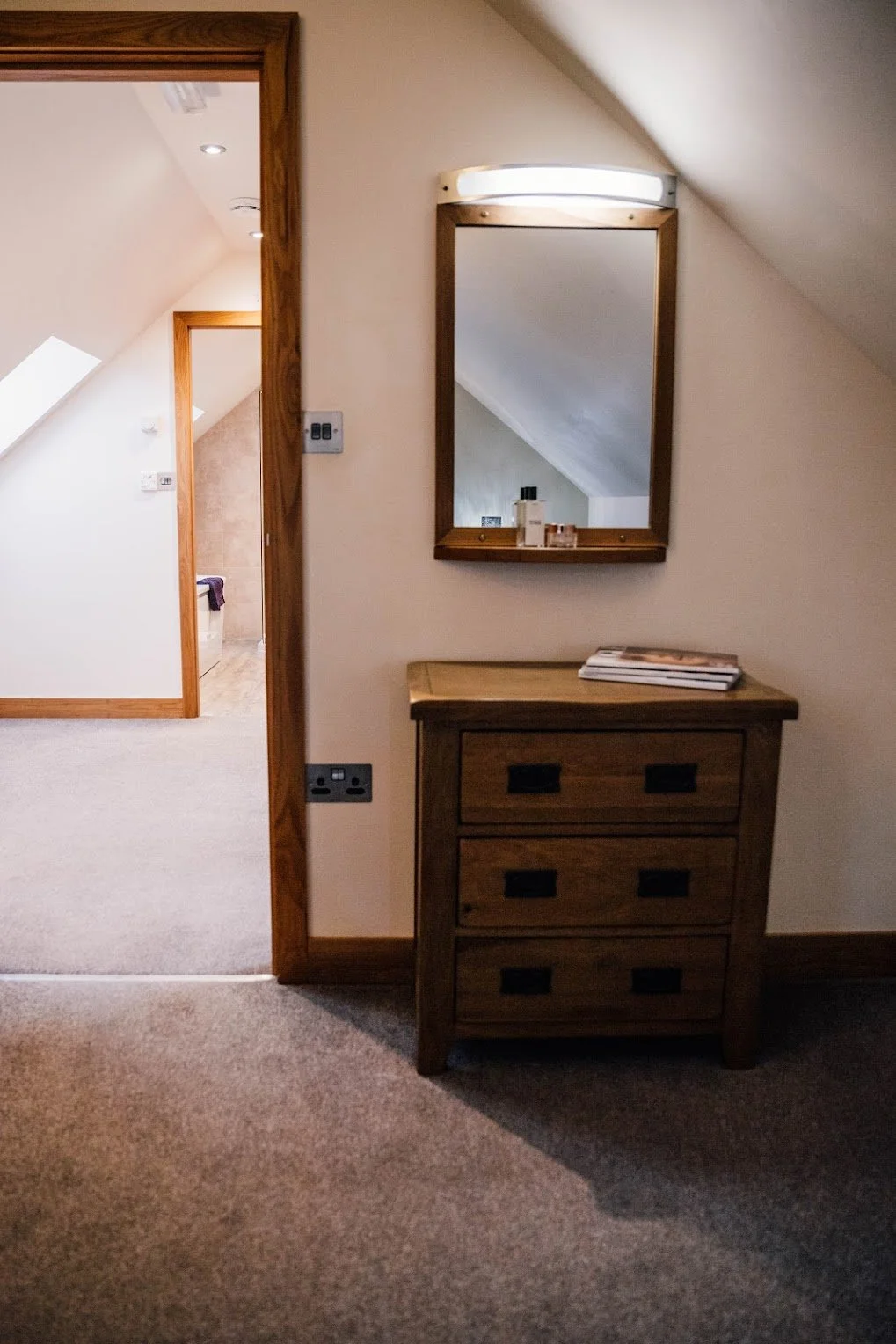 A wooden dresser with three drawers next to a wall mirror, small bottles and magazines on top, in a cozy room with beige carpeting and sloped ceiling.