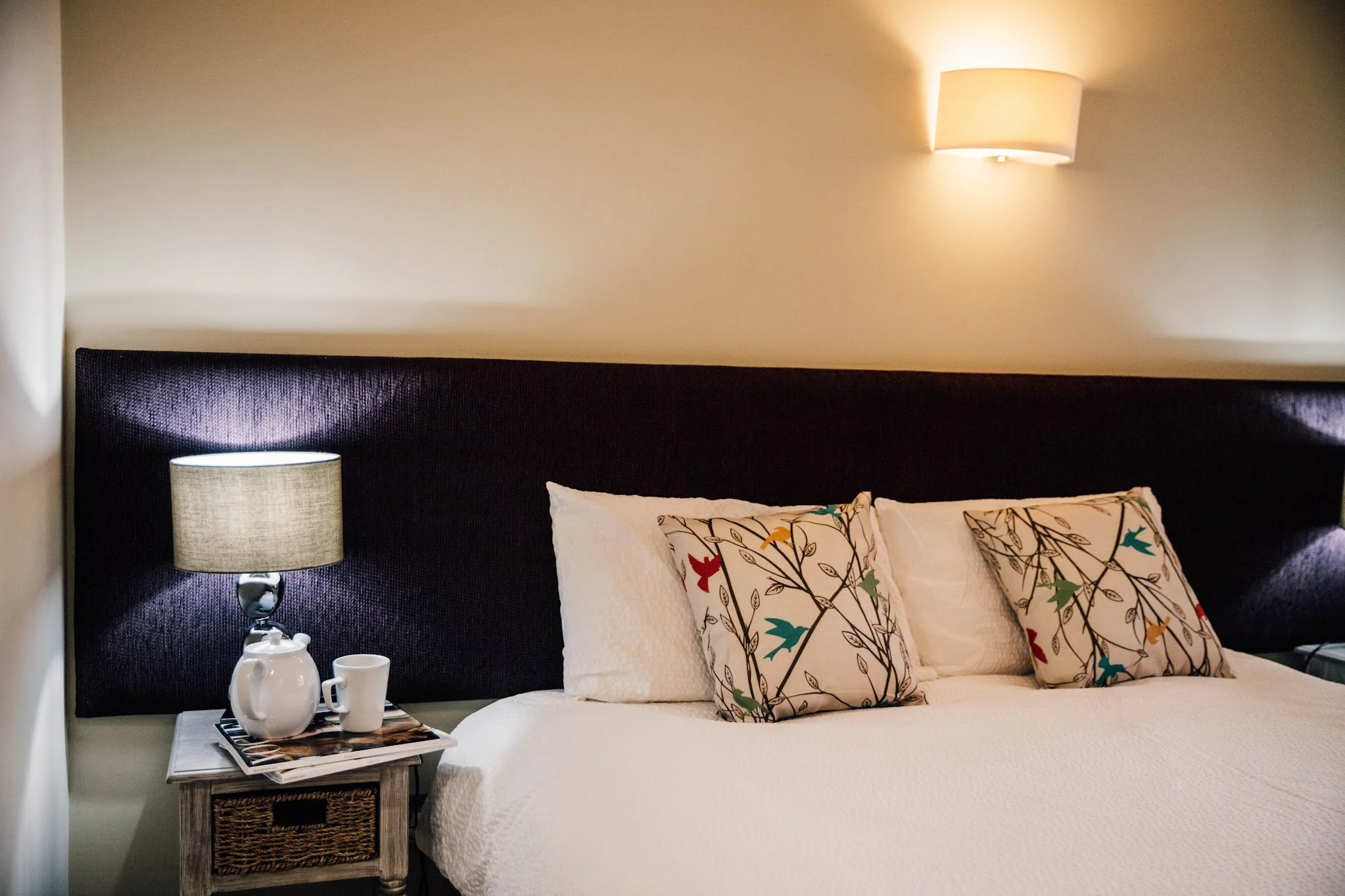 A cozy bedroom with a bed that has white bedding and decorative pillows with a floral design. A small wooden nightstand with a wicker basket drawer holds a white ceramic pitcher, a matching cup, and some magazines. A modern table lamp with a fabric shade sits on the nightstand. The wall behind the bed has a wall-mounted sconce light.