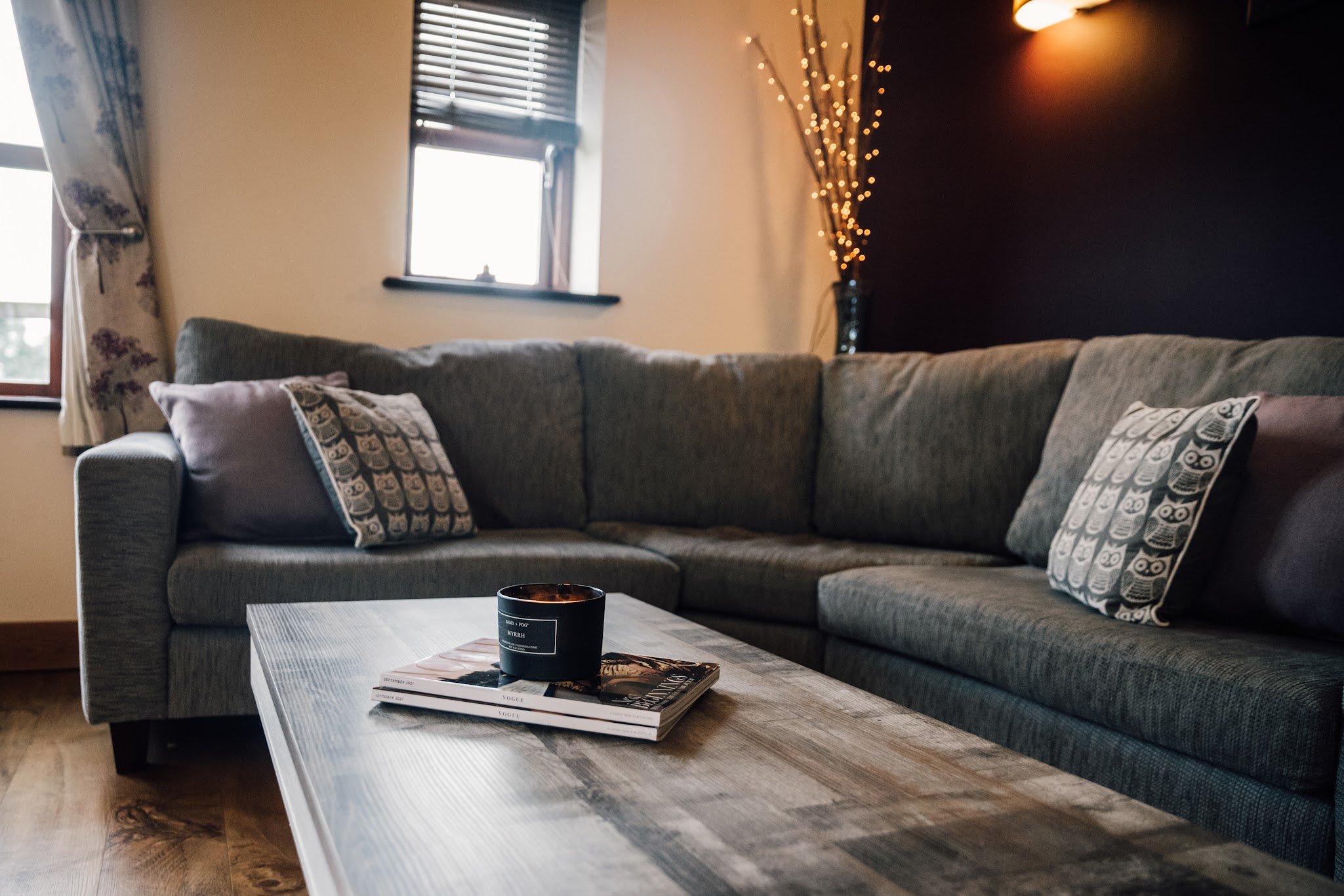 Living room with a grey sectional sofa and decorative cushions, a wooden coffee table with magazines and a candle, a window with curtains, and a dark accent wall with string lights in a vase.
