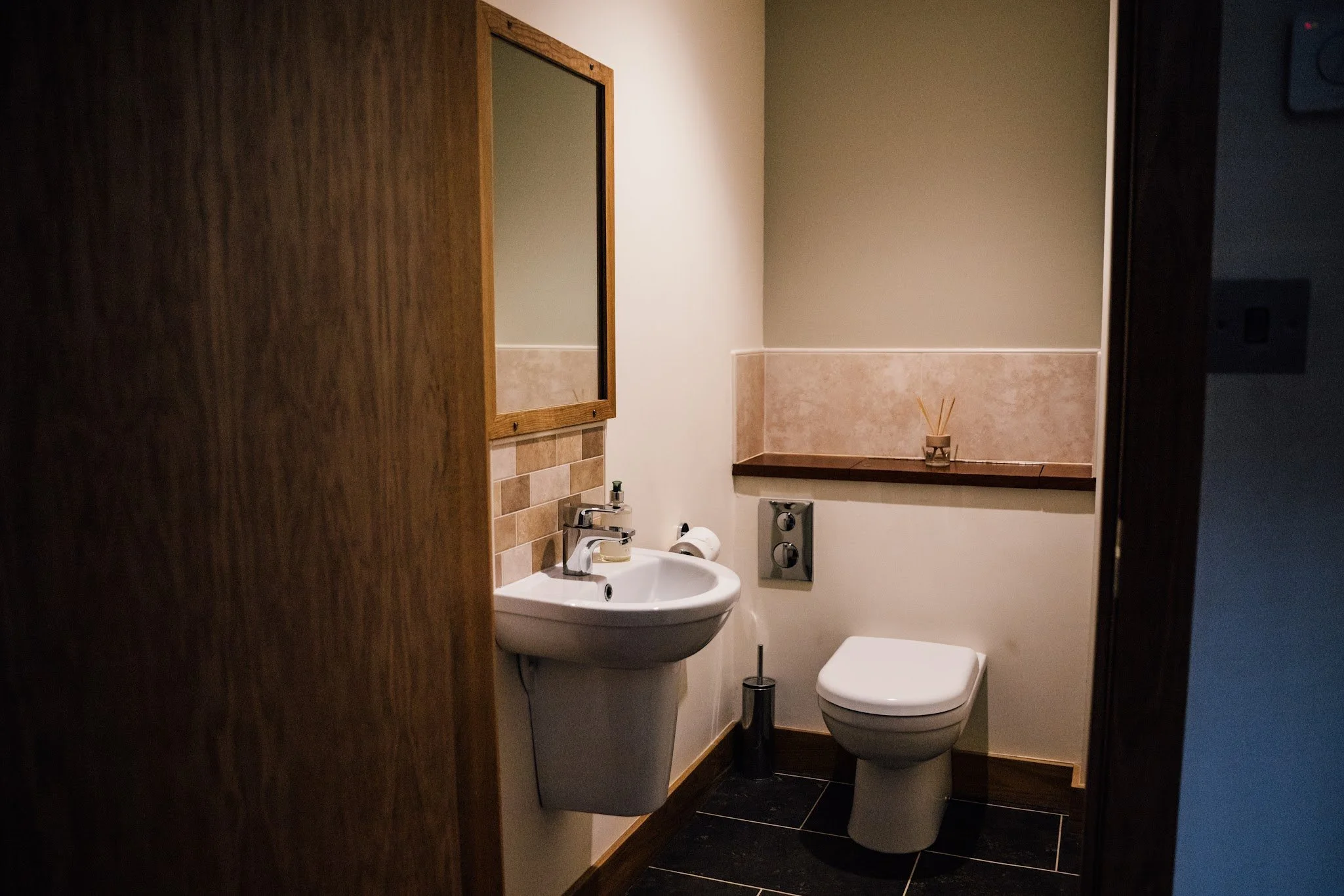 A small bathroom with a white sink, a mirror framed in wood, a toilet, and a small decorative item with reed diffusers on a wooden shelf. The walls are painted light green and have beige tile wainscoting. The floor is covered with black tiles.