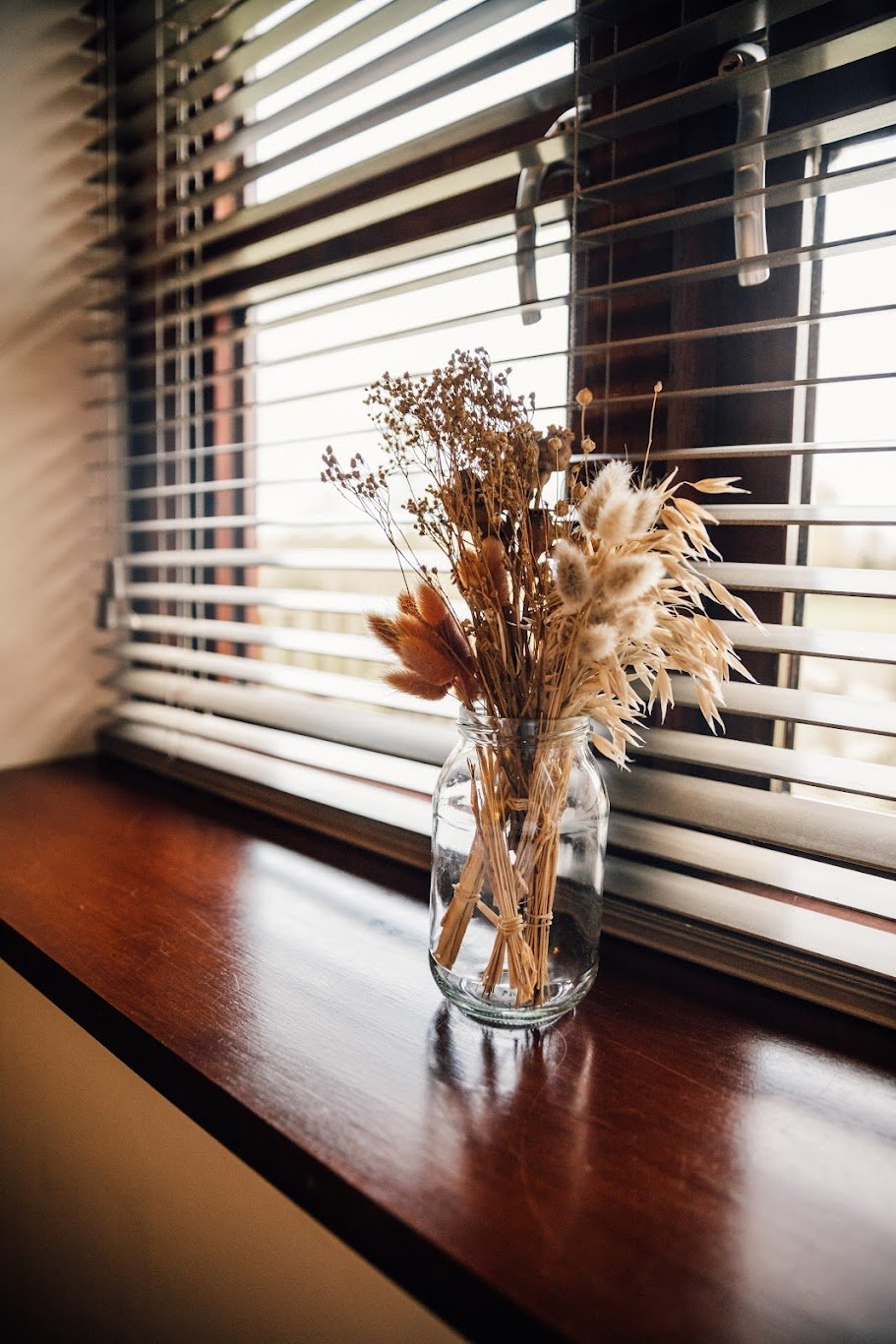 A clear glass jar filled with dried wildflowers and grasses sits on a wooden windowsill next to closed horizontal blinds allowing diffused light to enter.