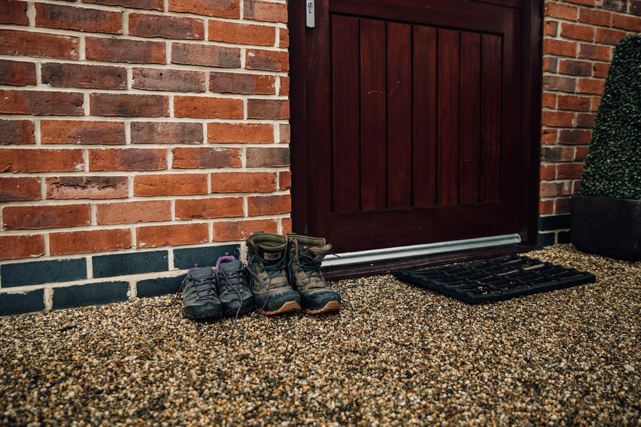 A brick wall with a dark wooden door, three pairs of hiking shoes on gravel outside the door, and gardening gloves on the gravel.