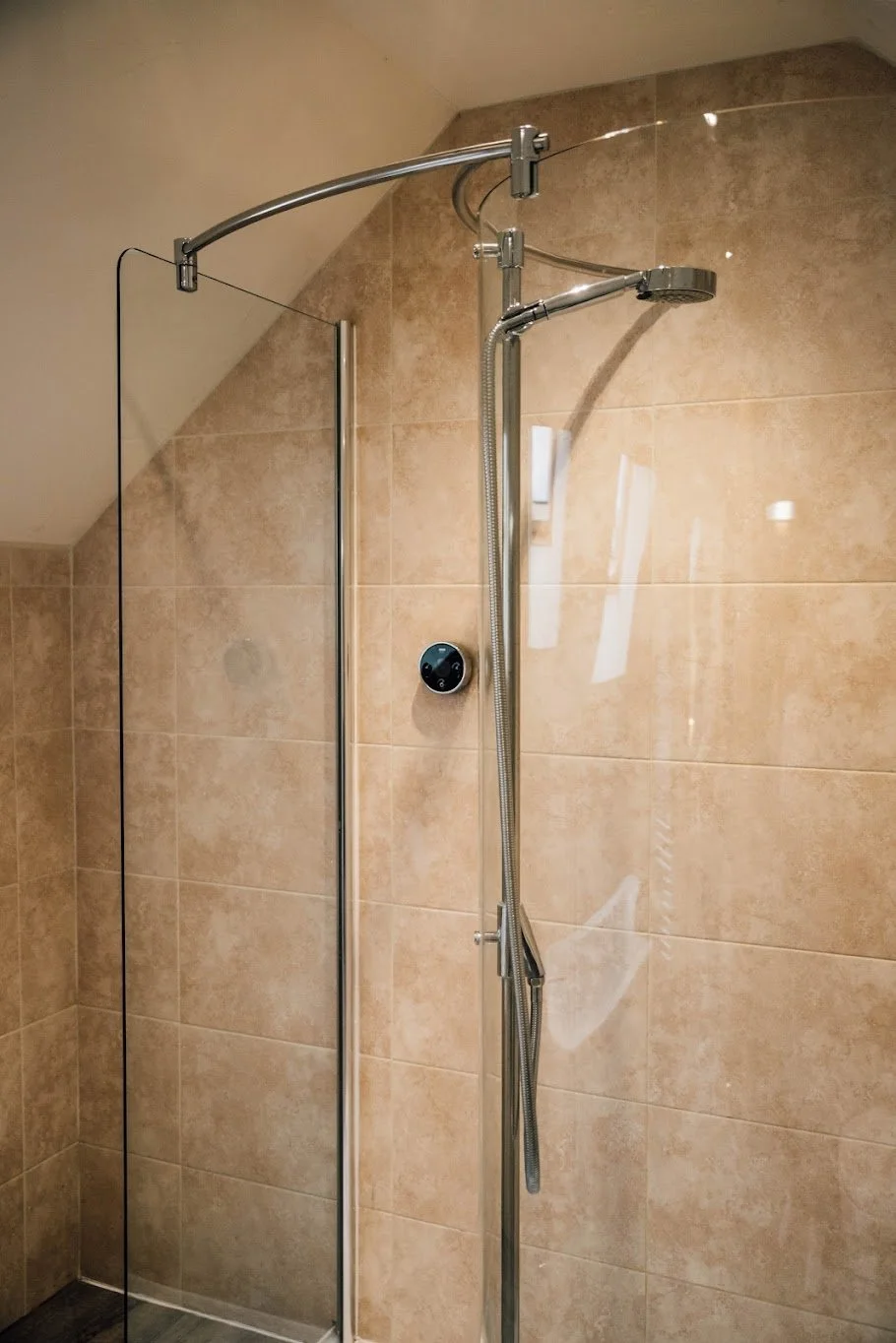A modern shower with beige tiles, a glass enclosure, a rainfall shower head, a handheld shower with a flexible hose, and a control knob.