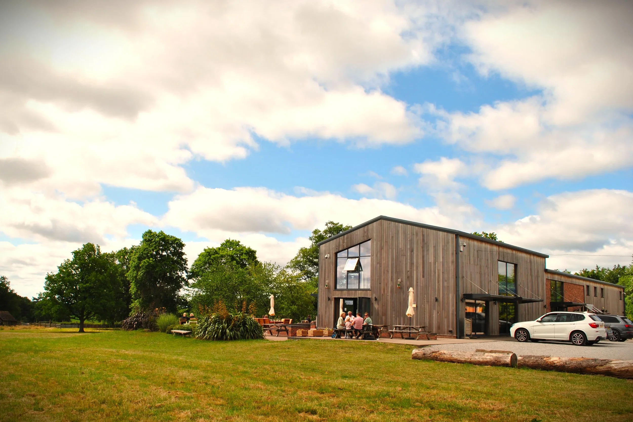 A modern wooden building with large windows in a park-like setting. There are people sitting at outdoor tables, some trees, and a cloudy sky overhead.
