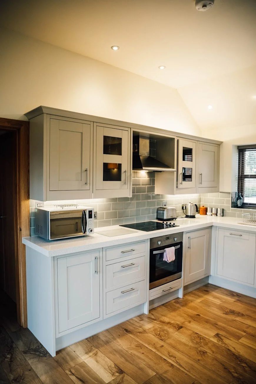 Modern kitchen with white cabinetry, stainless steel appliances, gray tile backsplash, wood flooring, and a window with blinds.