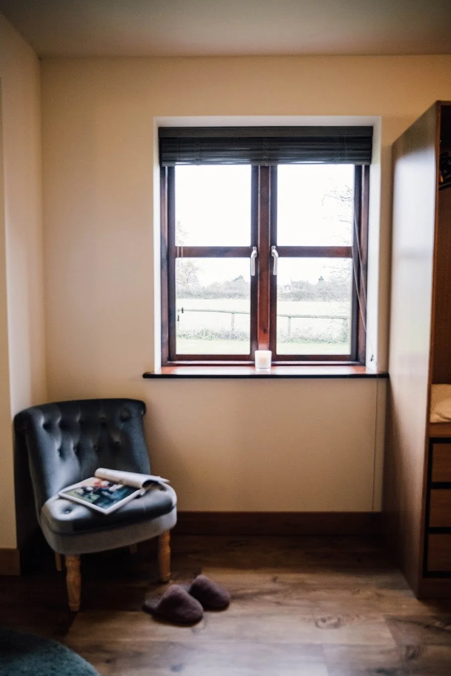 A cozy corner of a room with a window, a black tufted chair with magazines on it, brown slippers on the wooden floor, and a candle on the windowsill.