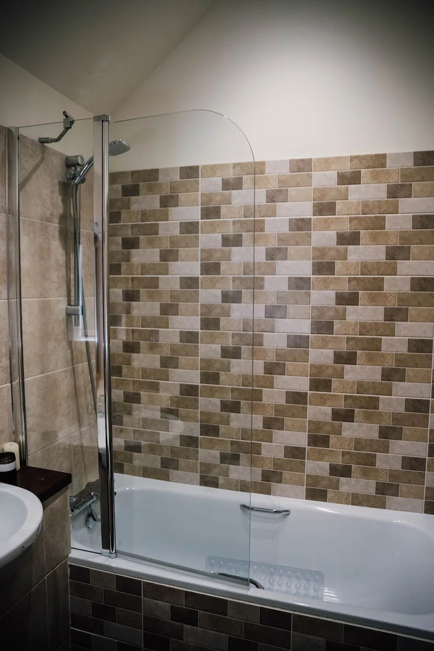 Bathroom with bathtub, glass shower door, beige and brown tiled wall, and a small section of a sink.