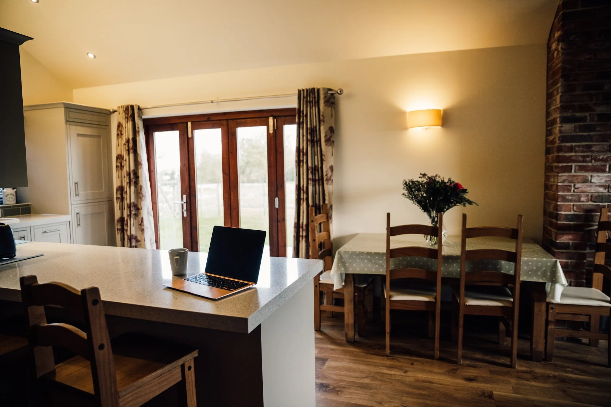 A kitchen and dining area with a large window, wooden chairs, a table with a vase of flowers, a laptop, a mug, and a coffee maker. The room has a brick wall and curtains.