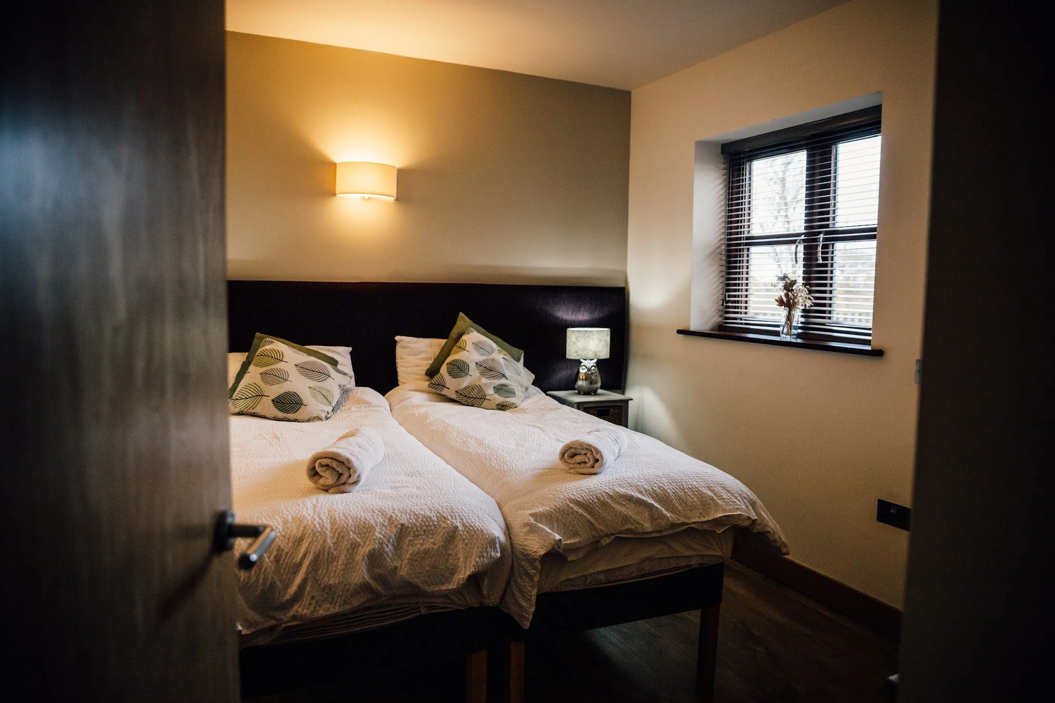 A cozy bedroom viewed through a partially open door. The bed has white bedding with decorative pillows, and two rolled towels are placed at the foot of the bed. On the right side, there is a window with blinds and a small table with a vase of dried flowers. A warm wall-mounted light fixture is on the wall above the bed, enhancing the inviting atmosphere.