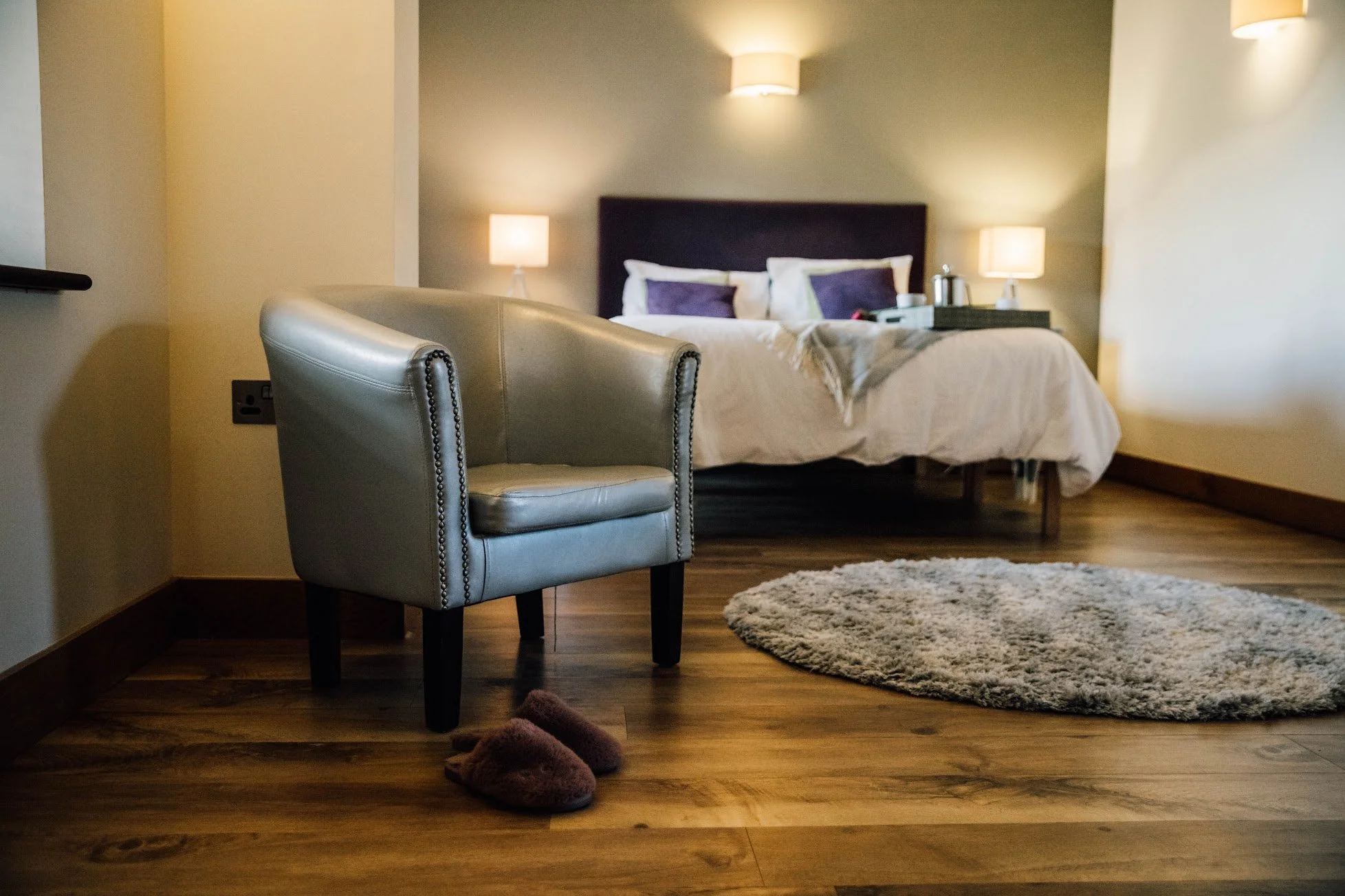 A bedroom with a bed, two bedside lamps, a chair in the foreground, a round rug, and slippers on a wooden floor.