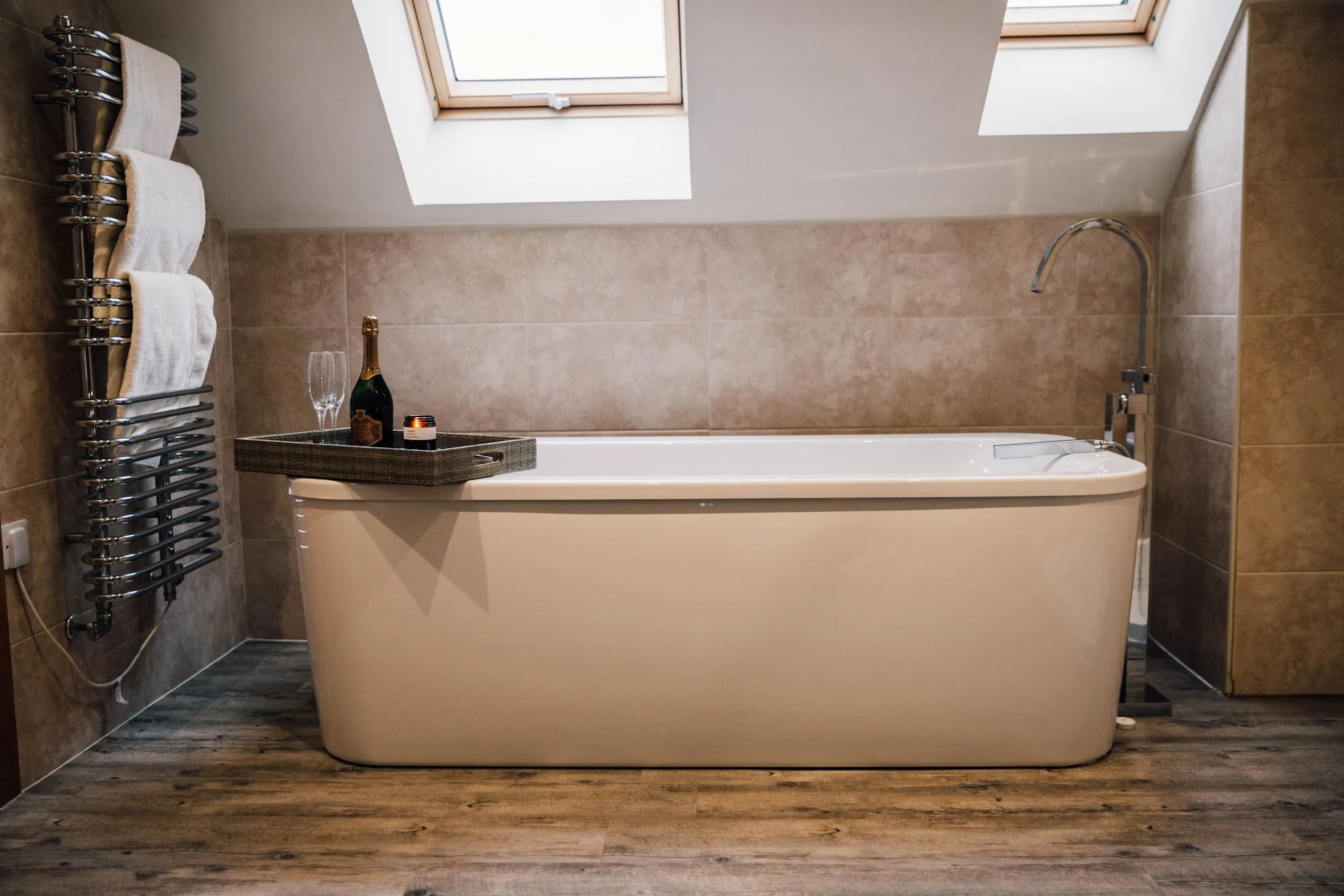 A bathroom with a bathtub beneath two skylight windows, a towel warmer with two folded towels, a tray with a bottle of champagne, two glasses, and a candle, with beige tiled walls and wooden flooring.