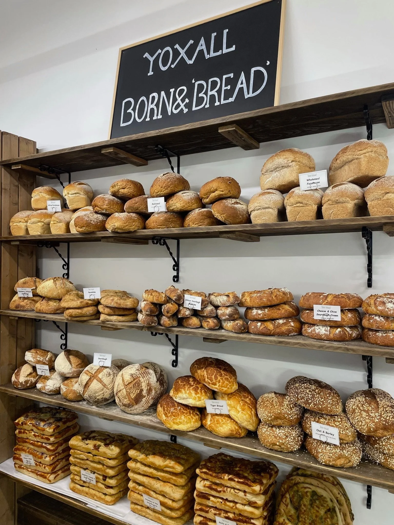 Display of various types of bread arranged on wooden shelves with labels, including sourdough, multiseed, oat & barley, and other specialty breads, with a sign above reading 'Yoxall Born & Bread.'