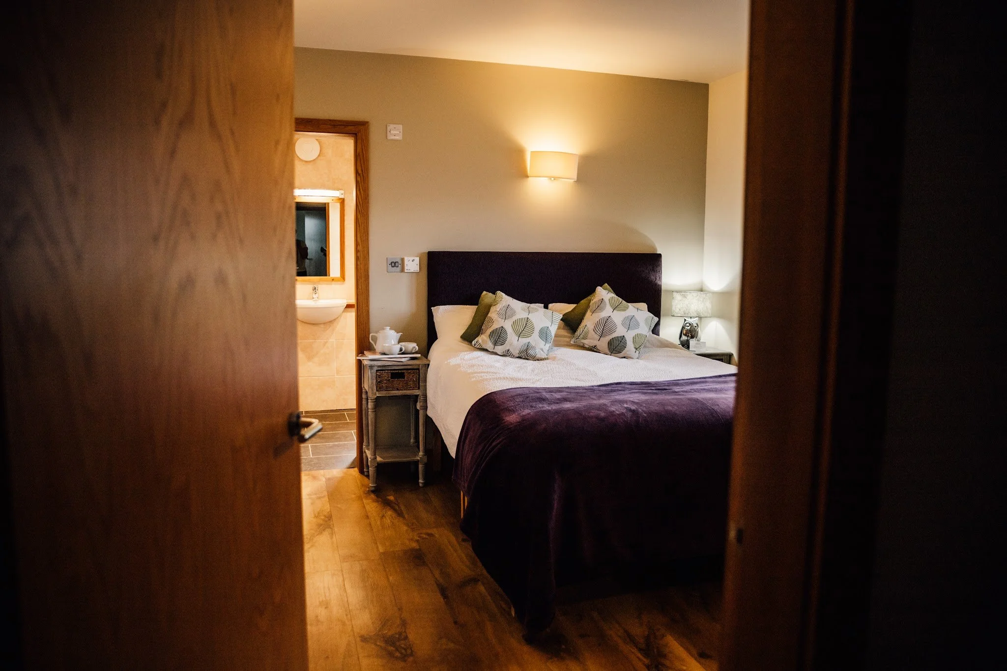 View of a neatly made bed with decorative pillows, a nightstand with a teapot and teacup, a wall-mounted lamp, and a window with a mirror in a cozy hotel room, seen through a partially opened wooden door.