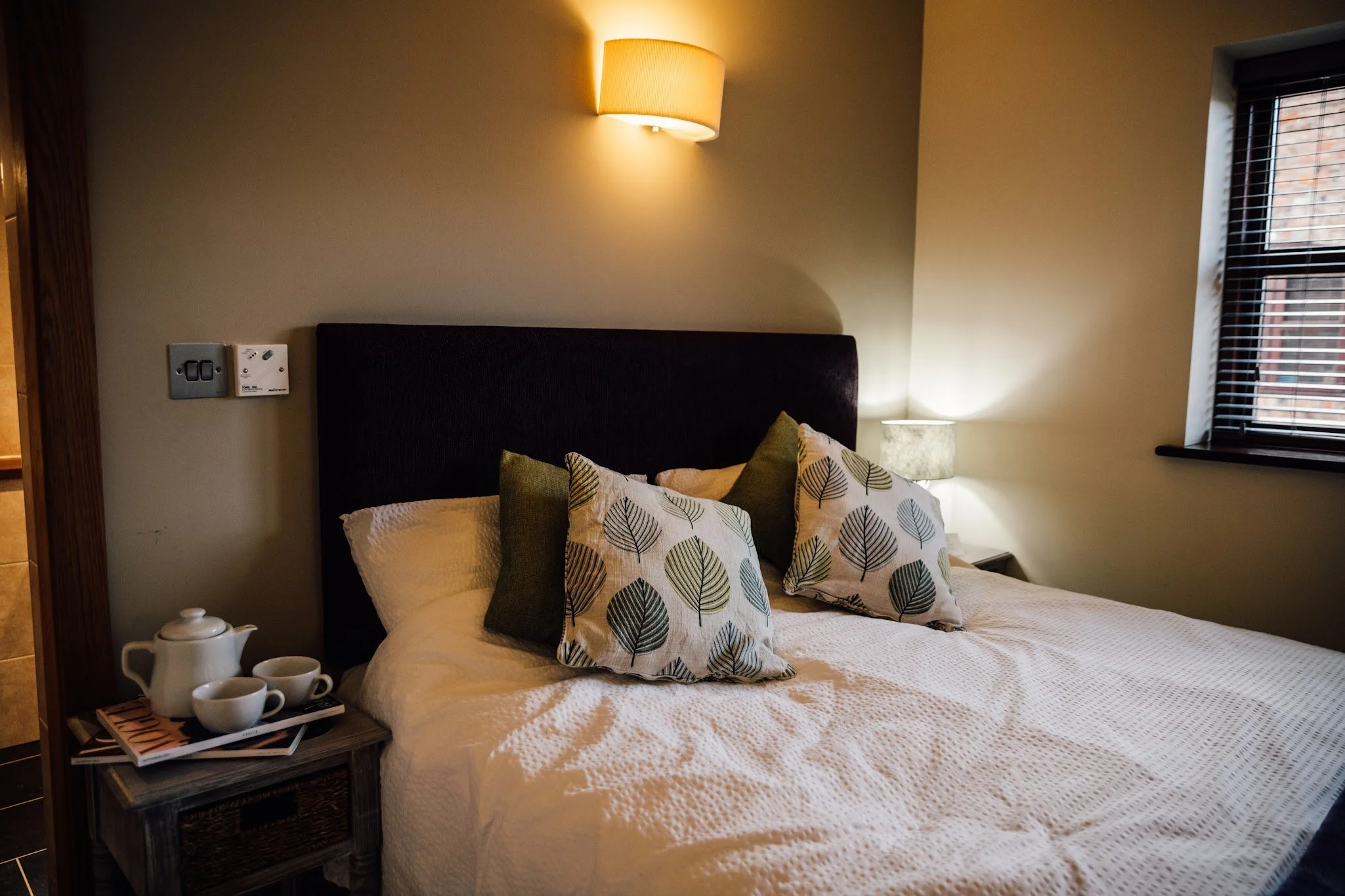 A bedroom with a bed, decorative pillows, wall-mounted lamp, nightstand with a tea set, window with blinds, and a wall switch.