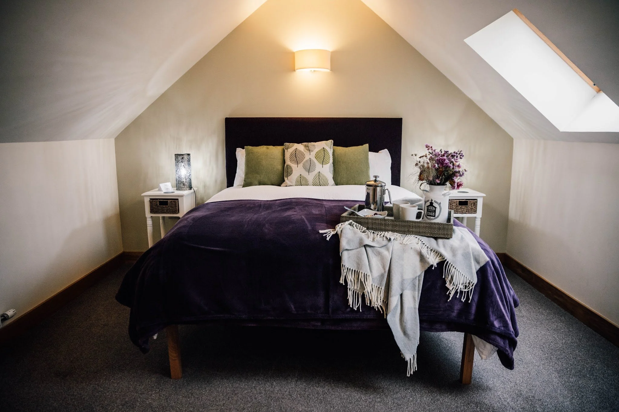A bedroom with a sloped ceiling, a bed with purple bedding, three pillows, nightstands on each side, a tray with a tea pot and cups, a flower vase, a lamp on the wall, and a skylight window.