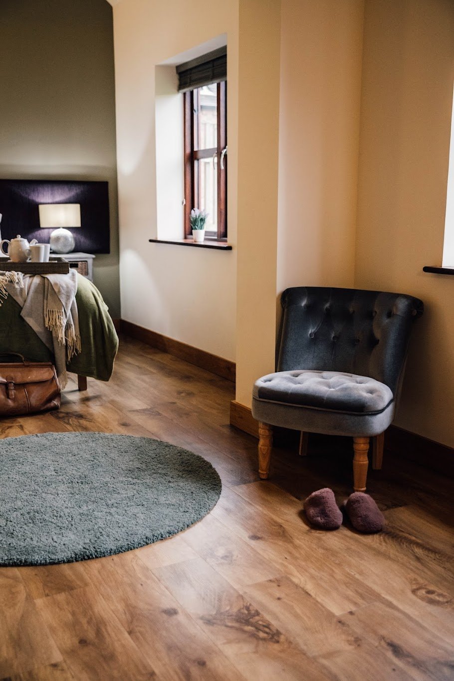 A cozy bedroom corner with a gray tufted armchair on wooden legs, pink slippers on hardwood floor, near a window with a small potted plant, and a partially visible bed with a bedside lamp and tea set.