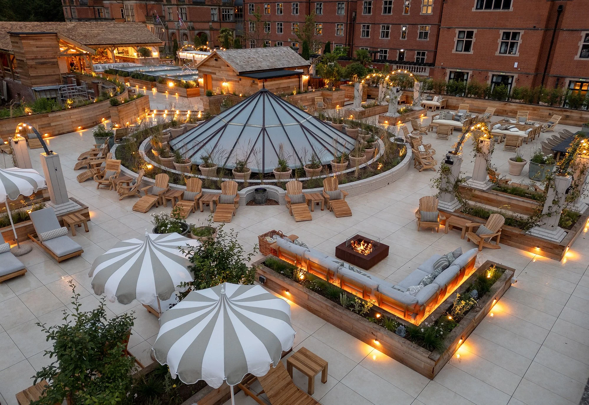 A rooftop terrace with outdoor furniture, umbrellas, plants, and string lights, overlooking a city block with brick buildings.