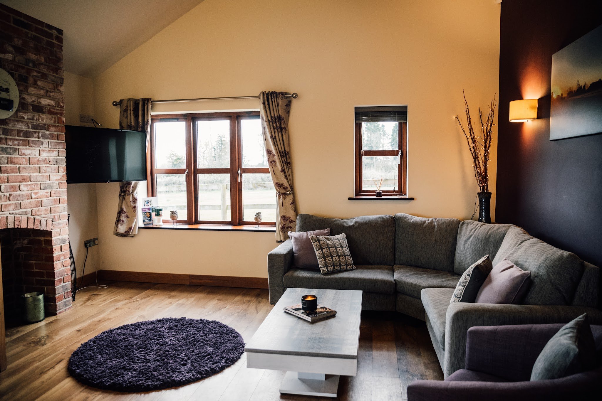 Living room with wooden flooring, a gray sectional sofa with patterned cushions, a white coffee table with magazines and a candle, a round purple rug, a brick fireplace, a large window with floral curtains, a smaller window, a wall-mounted TV, a decorative vase with branches, and a wall-mounted light
