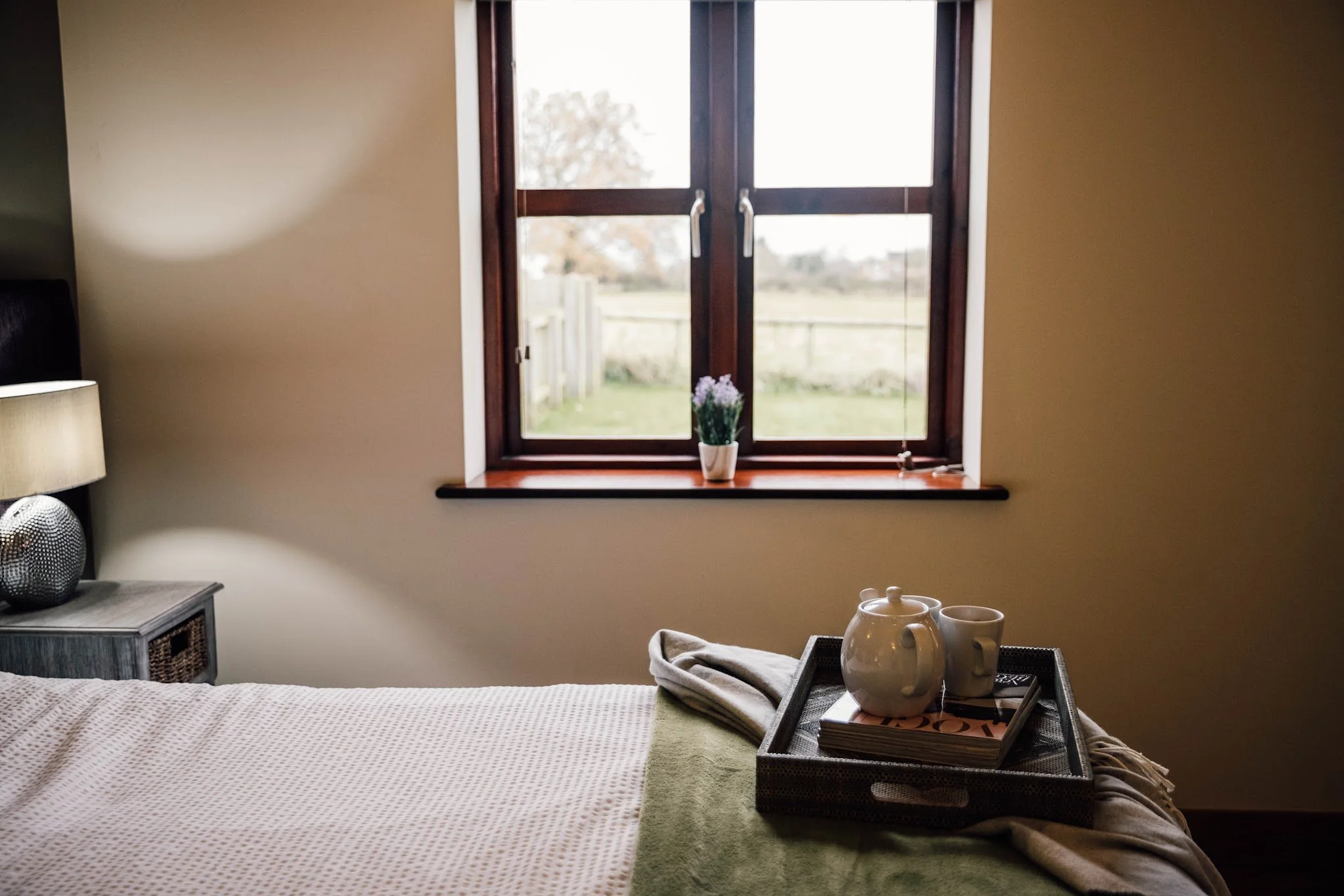 A bedroom with an open window, a bed with a green blanket, a nightstand with a lamp on it, and a tray with a white vase, cups, and magazines on the bed.