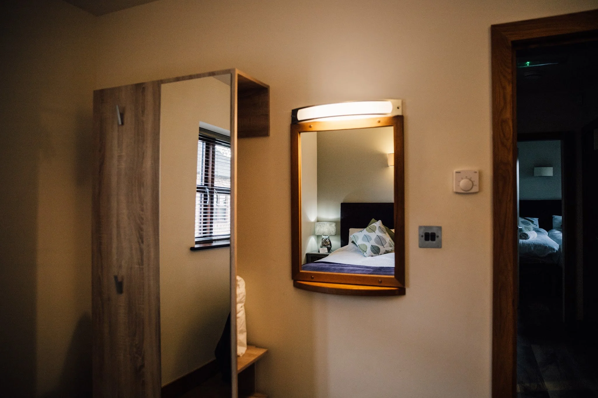 An interior view of a bedroom seen through a doorway with a large mirror on the wall reflecting the bed with patterned pillows and a nightstand with a lamp.